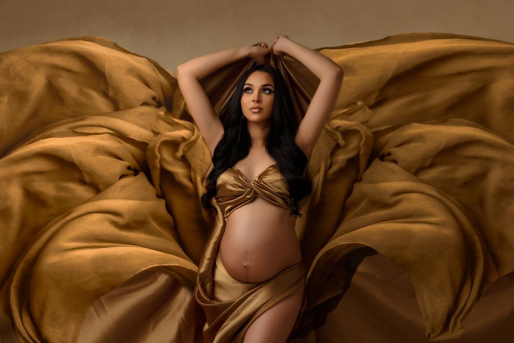 Dramatic maternity portrait with billowing golden fabric in moody lighting.