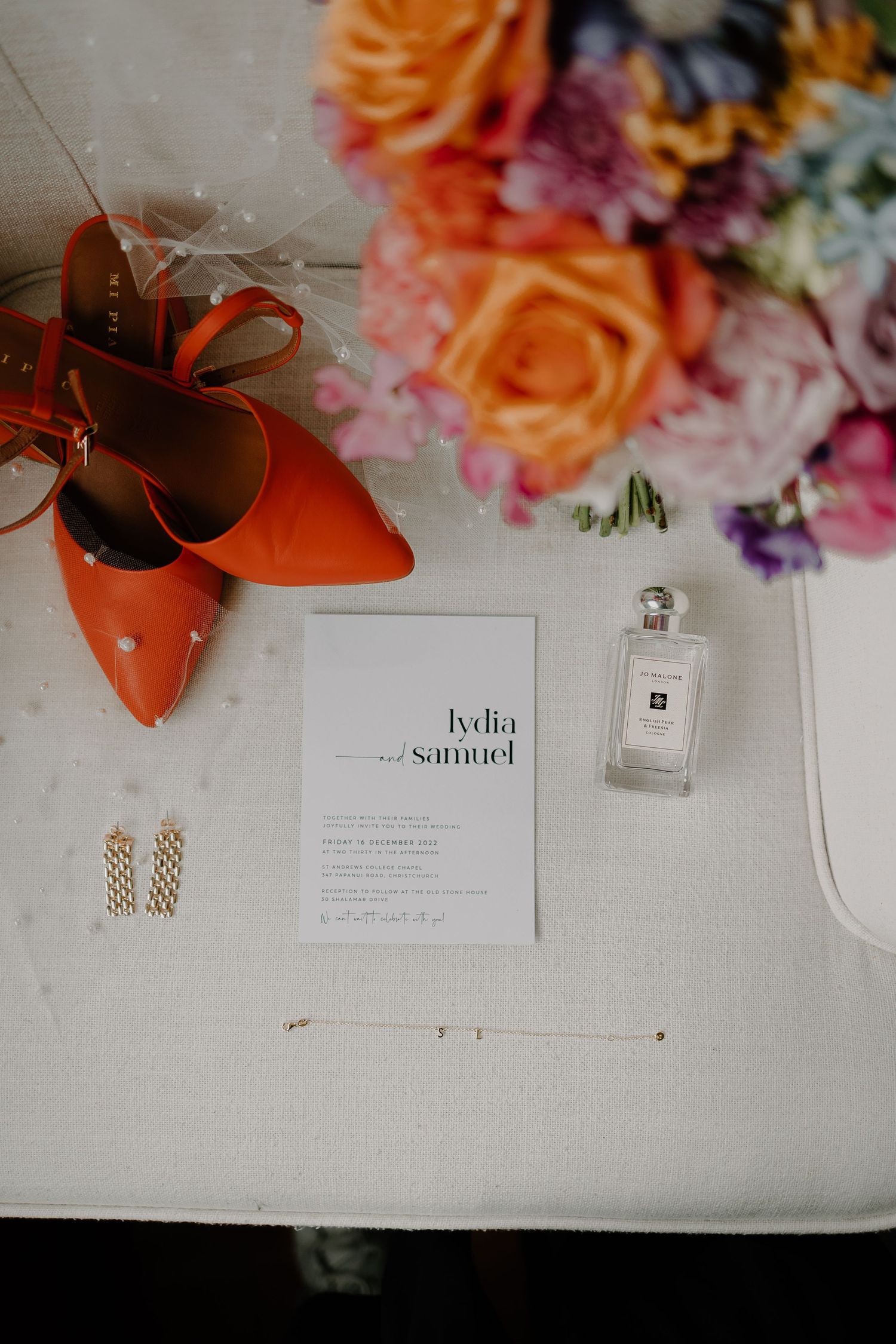 Wedding details flatlay with orange shoes, invitation, perfume bottle and colorful roses.