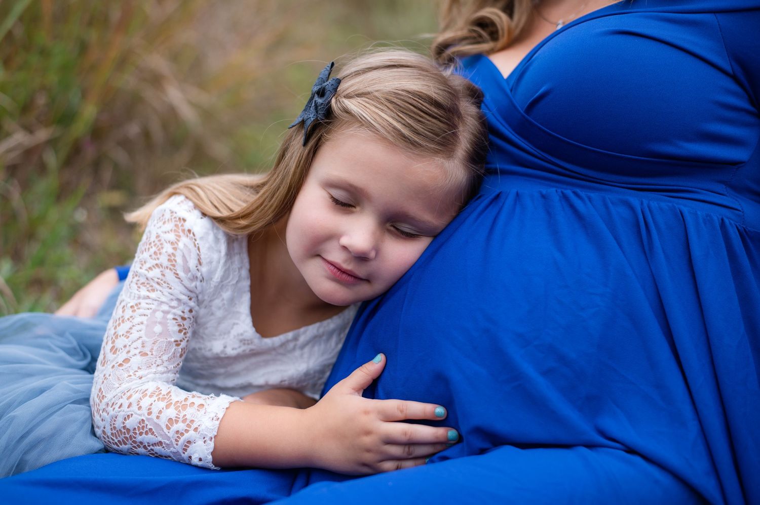 Sweet moment captured as young girl rests head on mother's pregnant belly in outdoor setting.