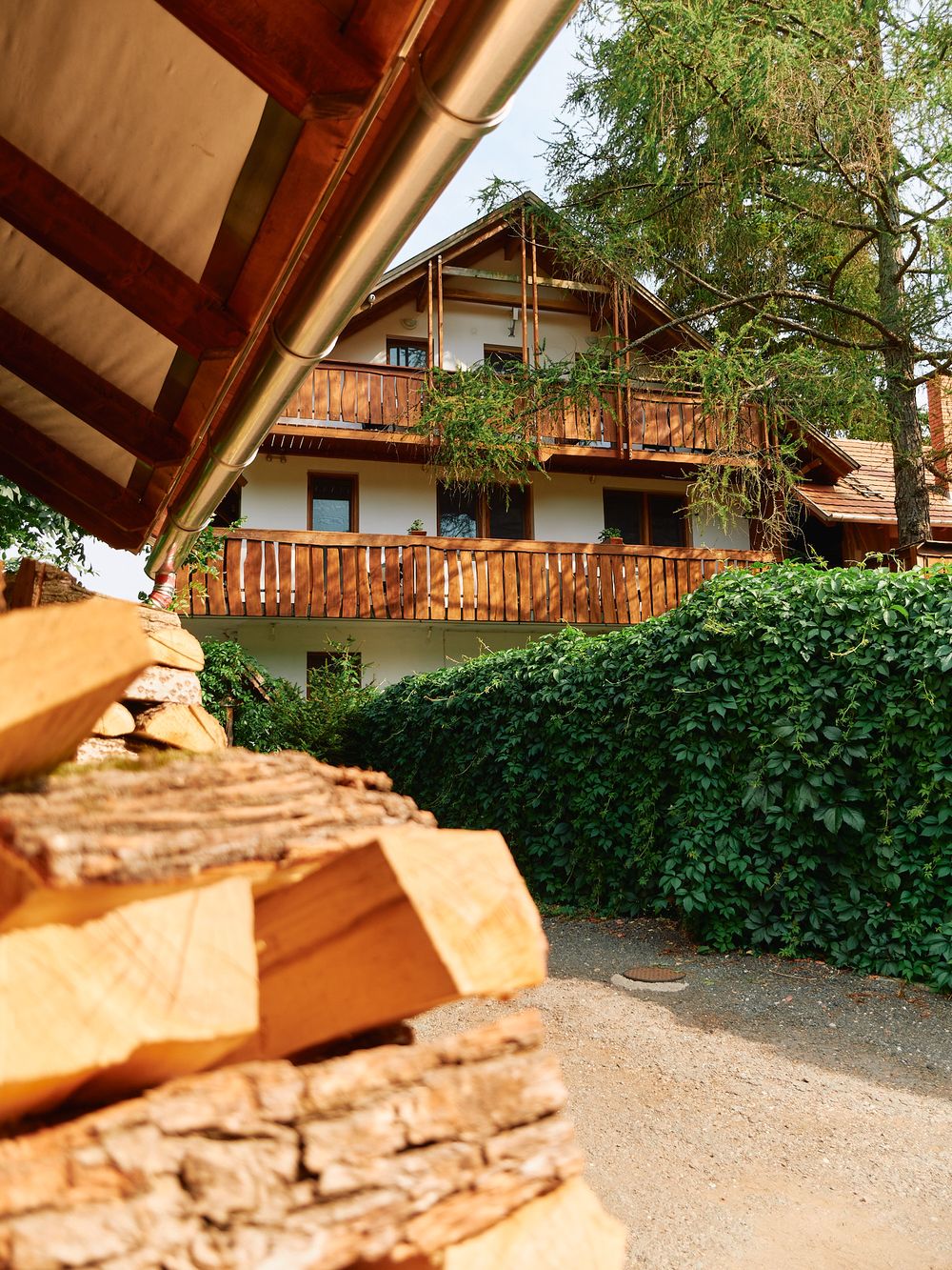 Alpine-style wooden house with balconies viewed through stone wall and greenery.