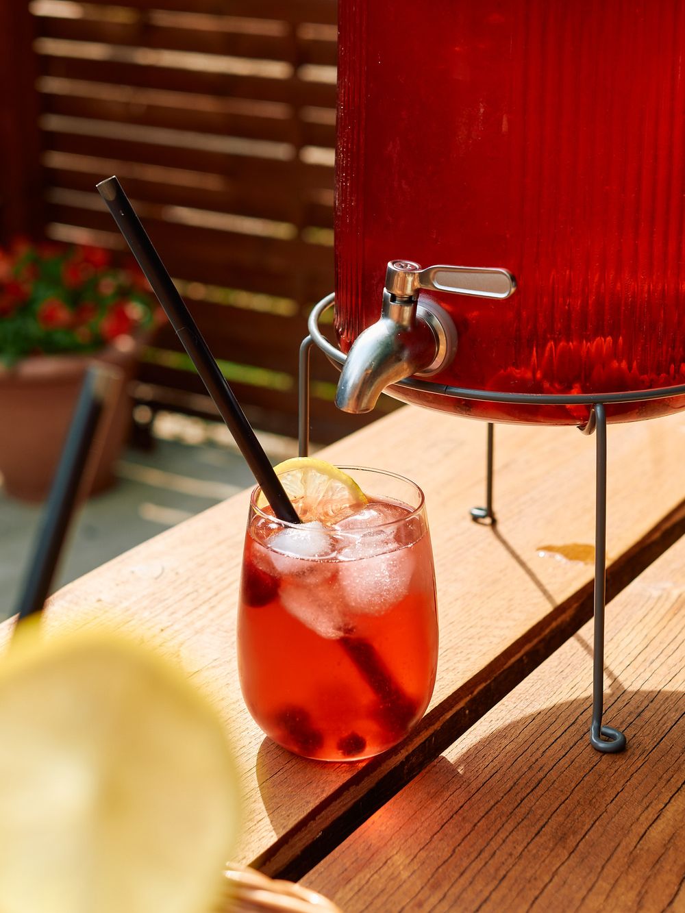 Red refreshing cocktail with ice in glass beside beverage dispenser on wooden surface.