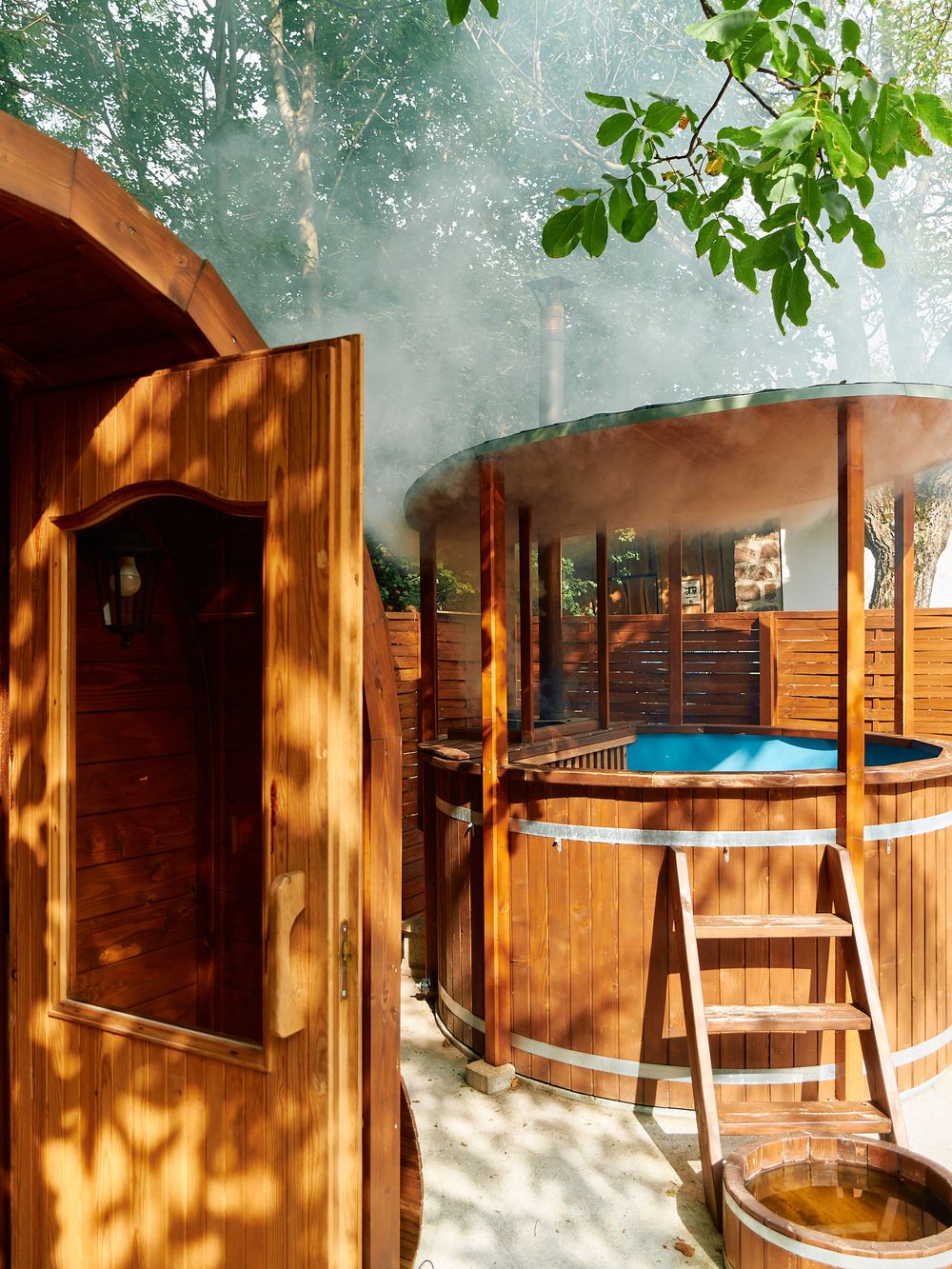 Wooden hot tub and sauna setup in outdoor garden setting with dappled sunlight.