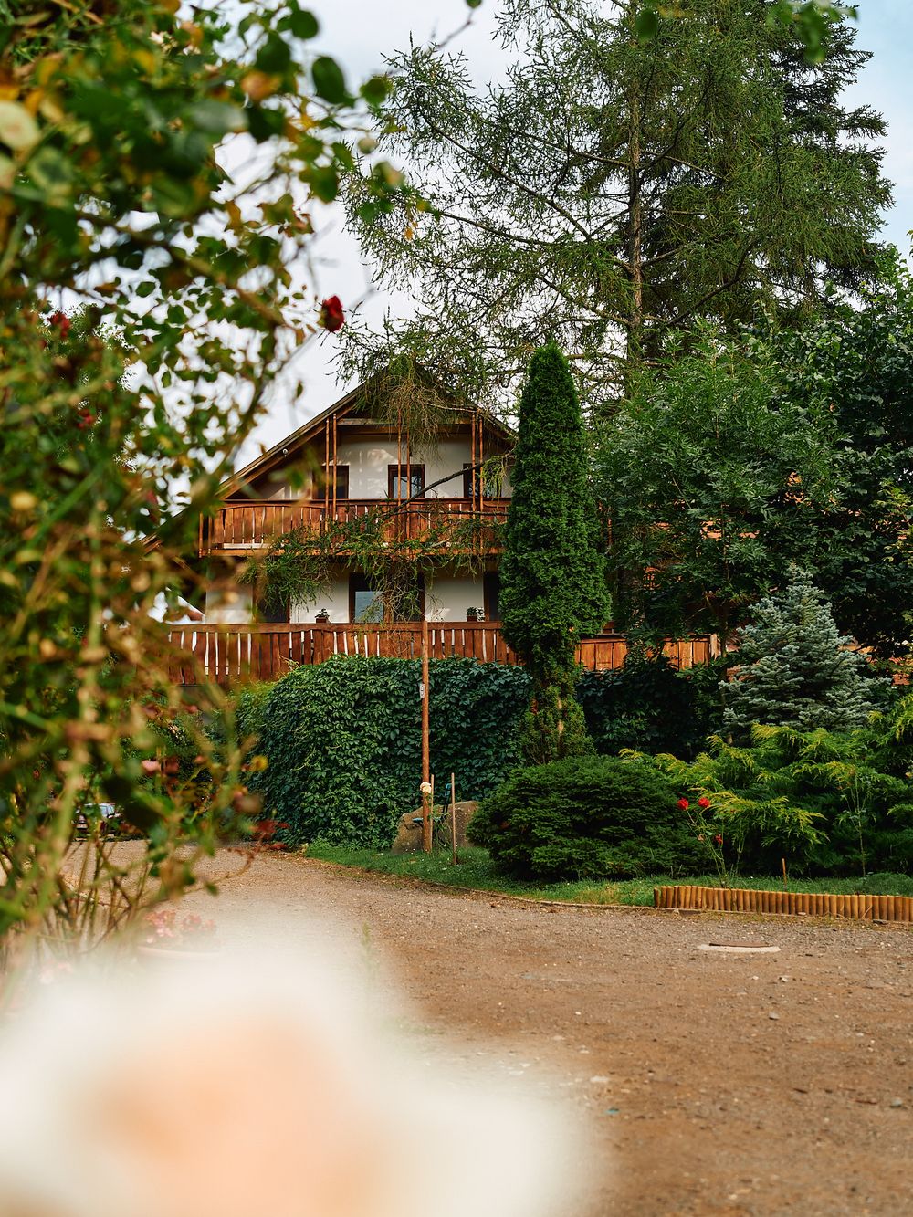 Two-story alpine style house with wooden balconies surrounded by lush greenery.