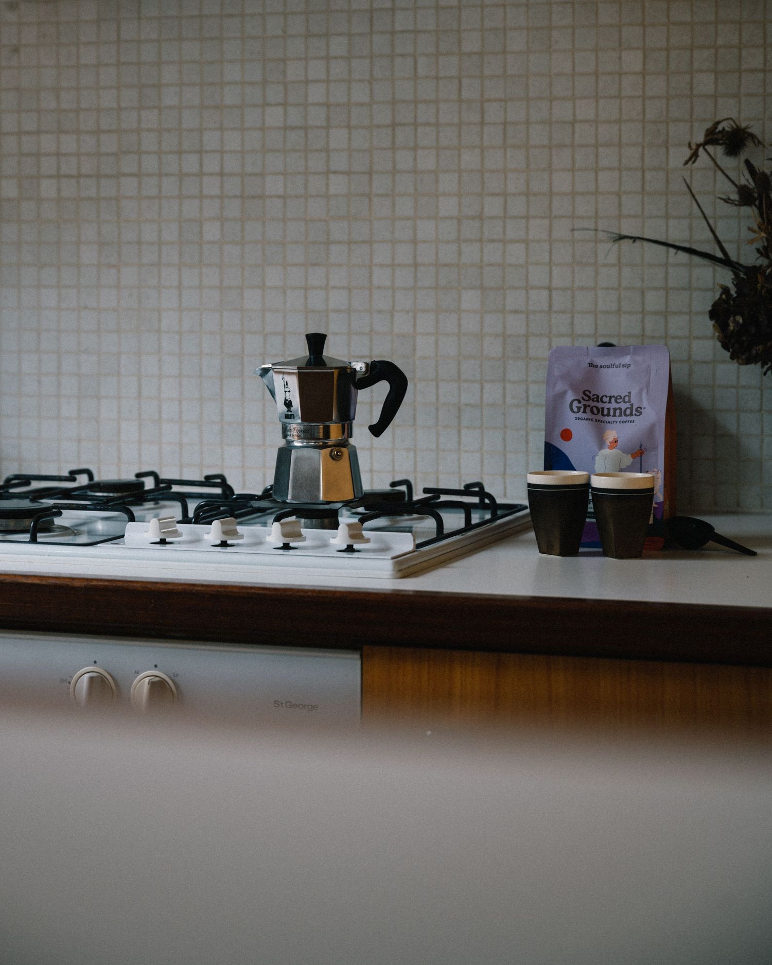 Stovetop with moka pot coffee maker and coffee grounds on white tiled kitchen counter.