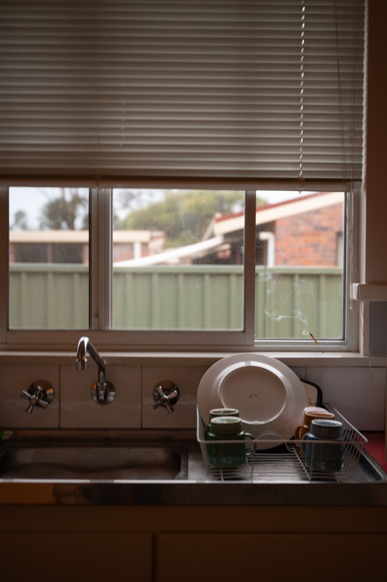Vintage kitchen sink with white blinds and window overlooking suburban backyard.