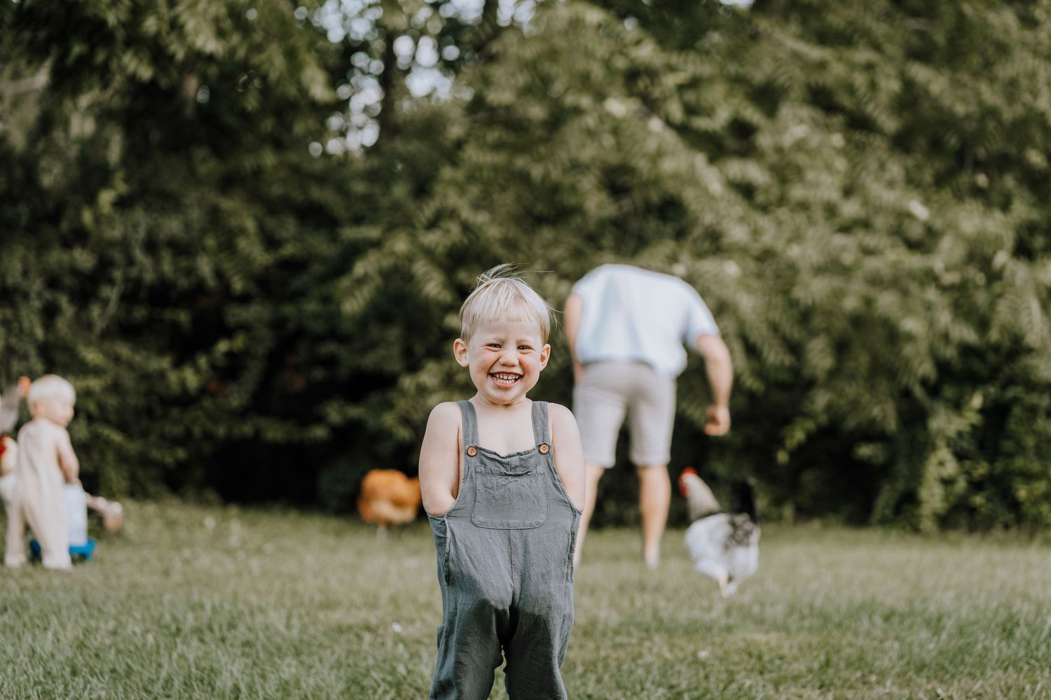 Young child in overalls smiling brightly while playing in a sunny backyard.