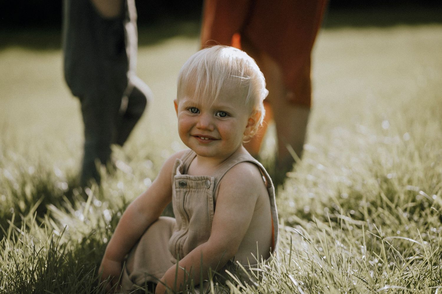Toddler in overalls sitting in sunlit grass with a happy expression.