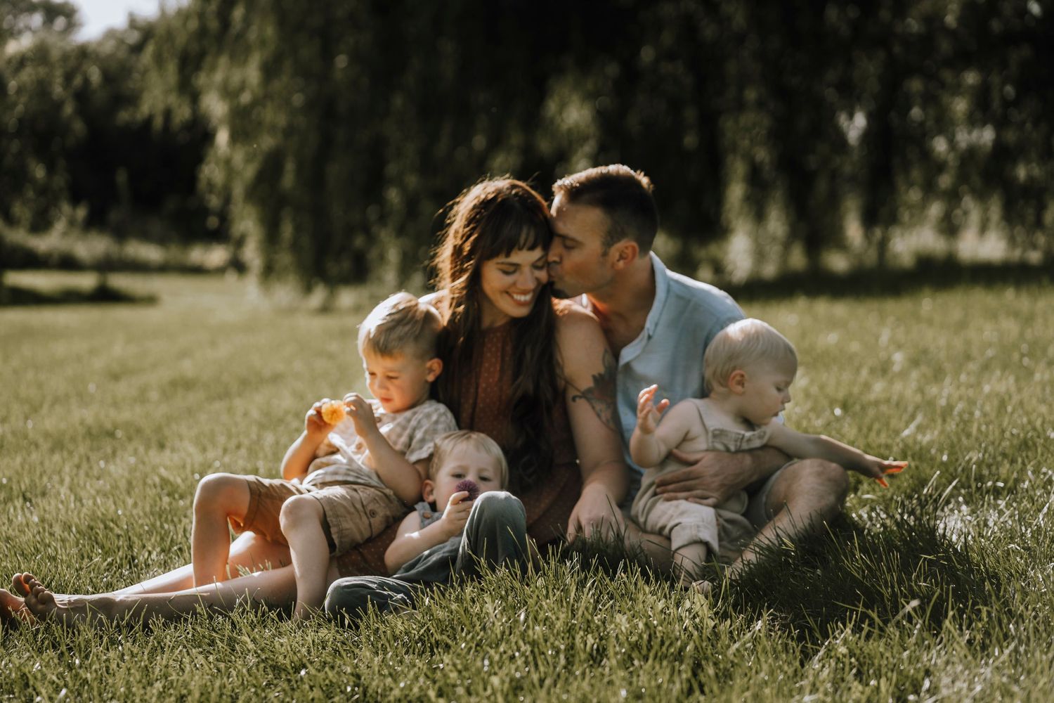 Family enjoying tender moment together in sunny park with willow trees.
