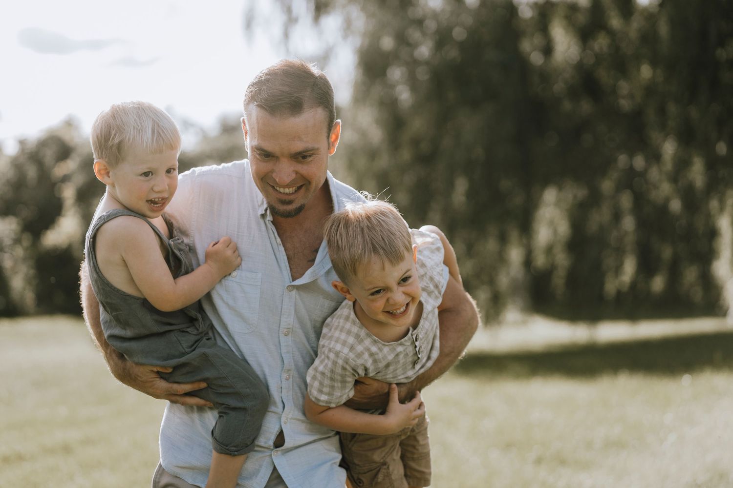 A loving family moment outdoors as a parent holds two small children in a warm summer setting.