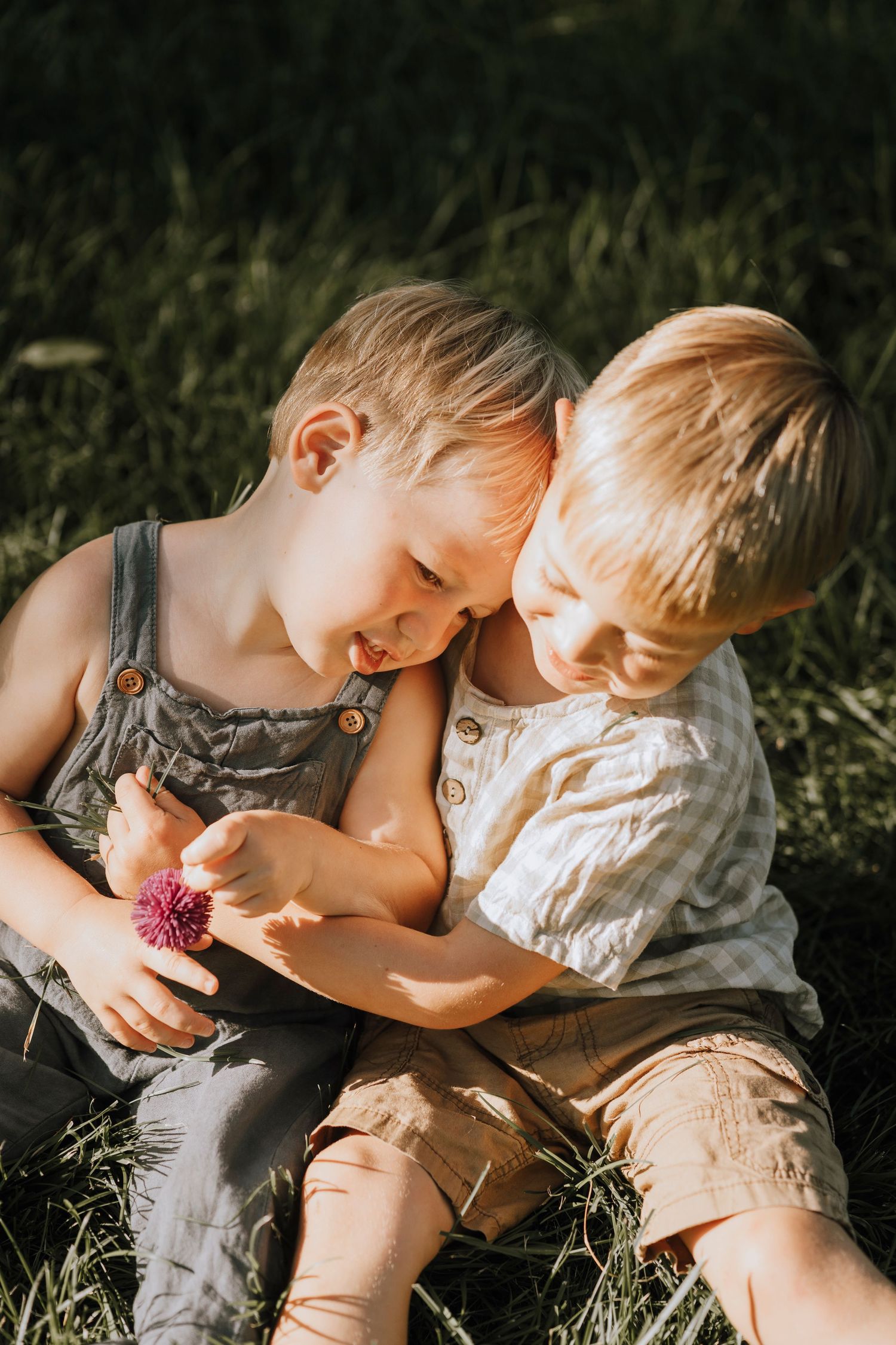 Two young children sitting close together in overalls examining something in the grass.