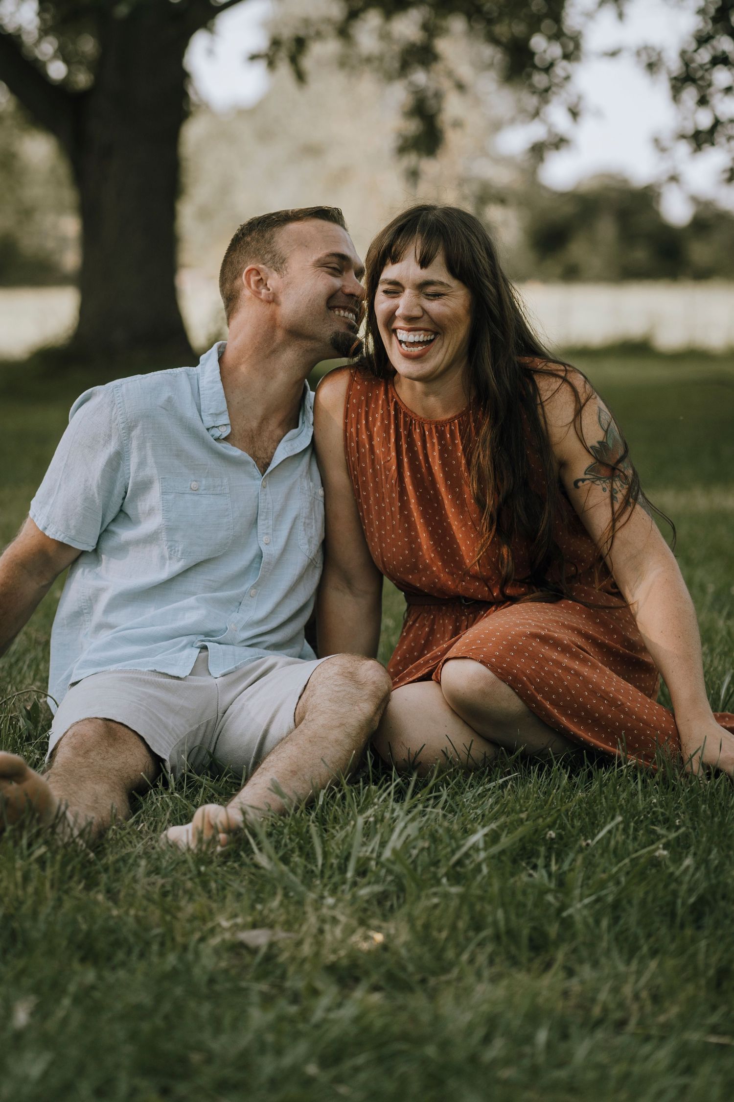 Two people share genuine laughter while sitting together on grass in a park.