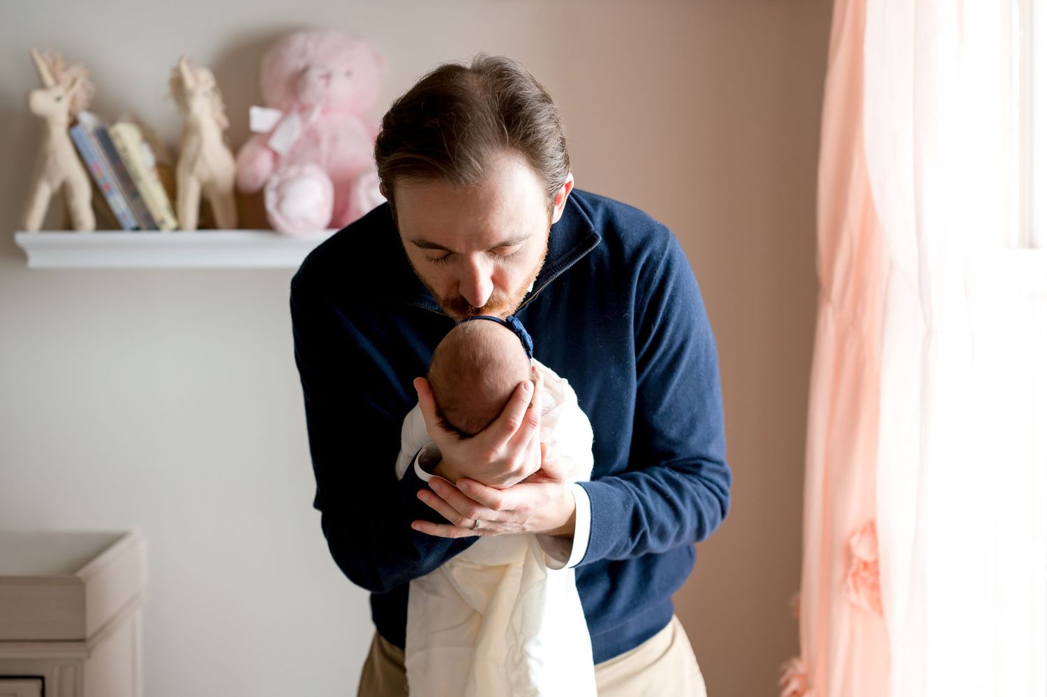 father wearing a blue sweater kissing the forehead of his newborn baby girl inside her pink soft nursery in west u