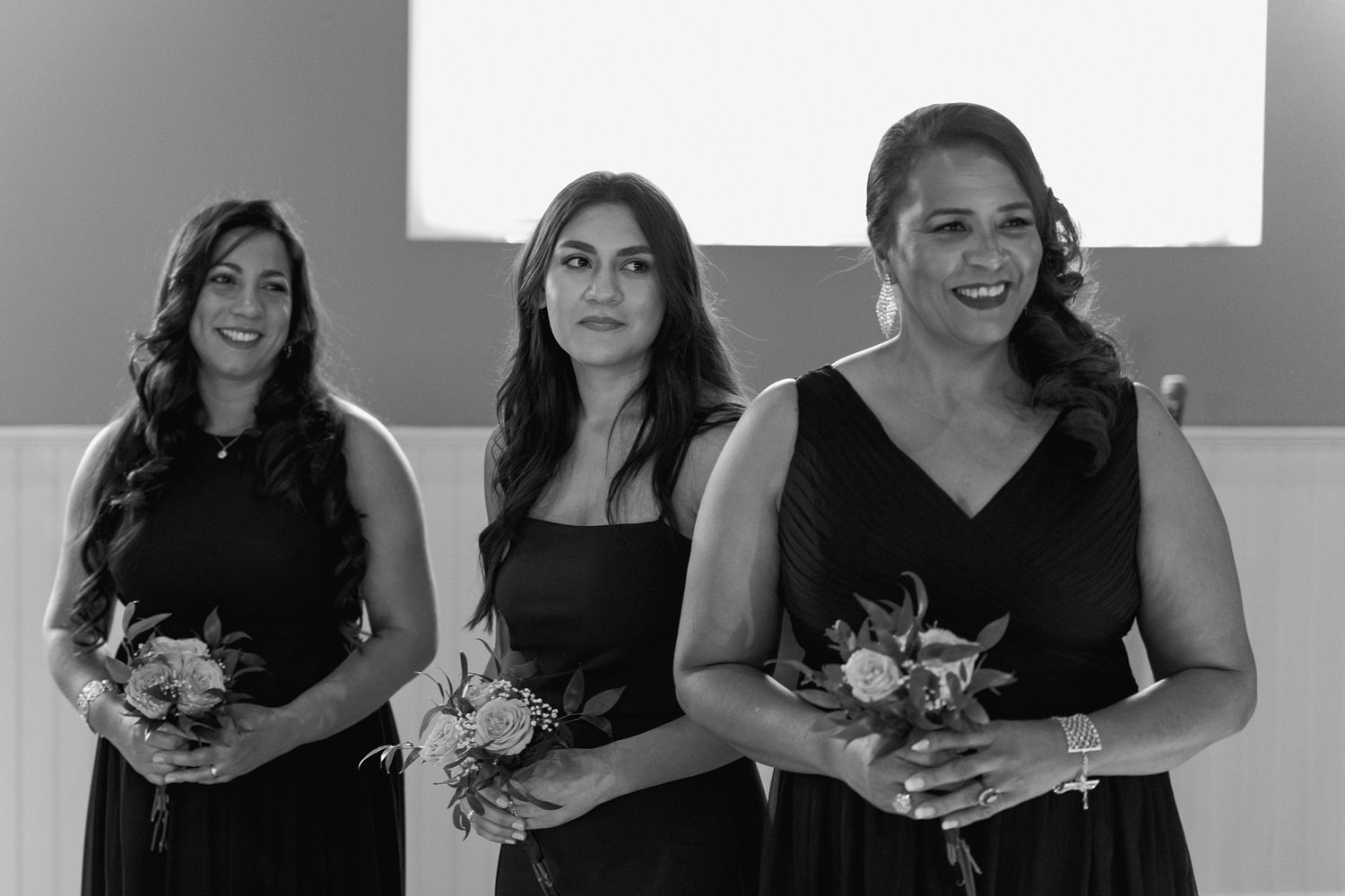 Three bridesmaids in black dresses hold bouquets while smiling.