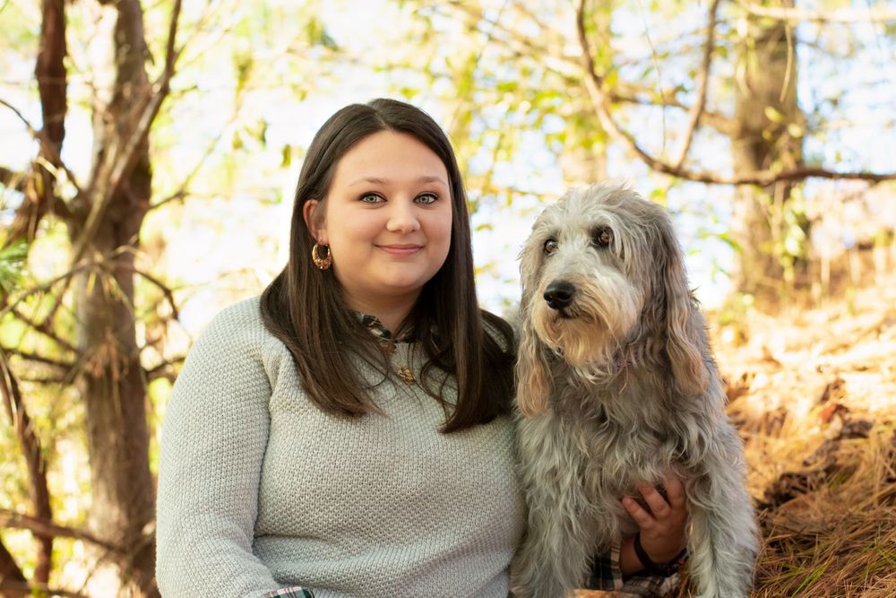A grey shaggy dog and person sit among fallen autumn leaves in a wooded setting.
