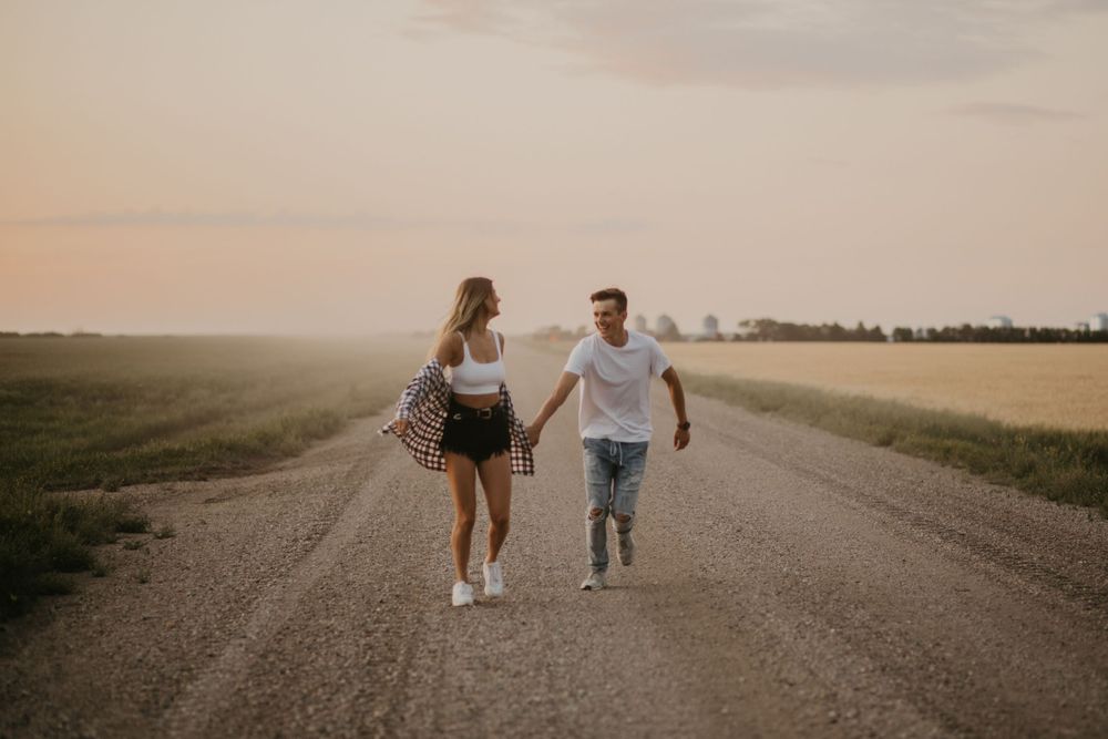 A couple runs playfully down a dirt road at sunset through rural farmland.