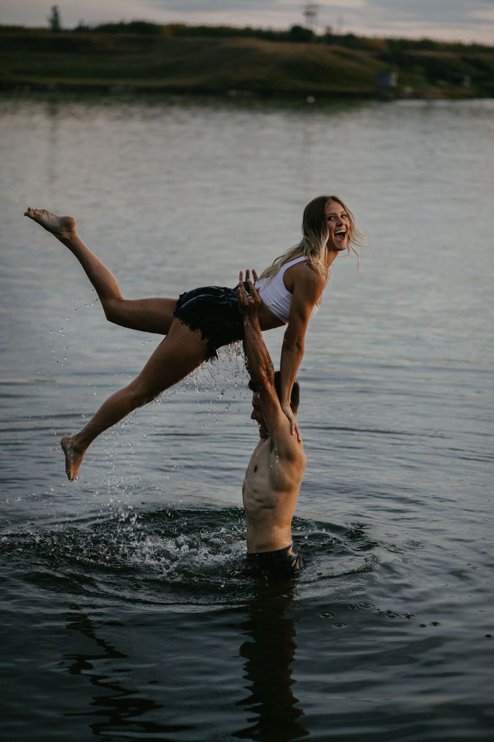 A playful couple performs an acrobatic lift pose in a lake during sunset.