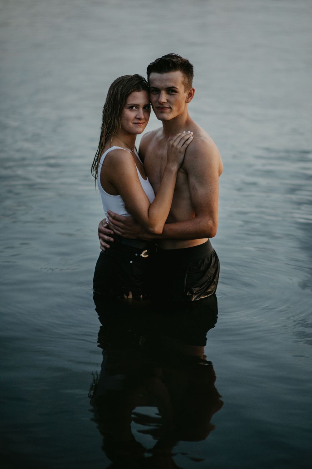 A romantic couple poses together while standing in a peaceful lake during sunset.