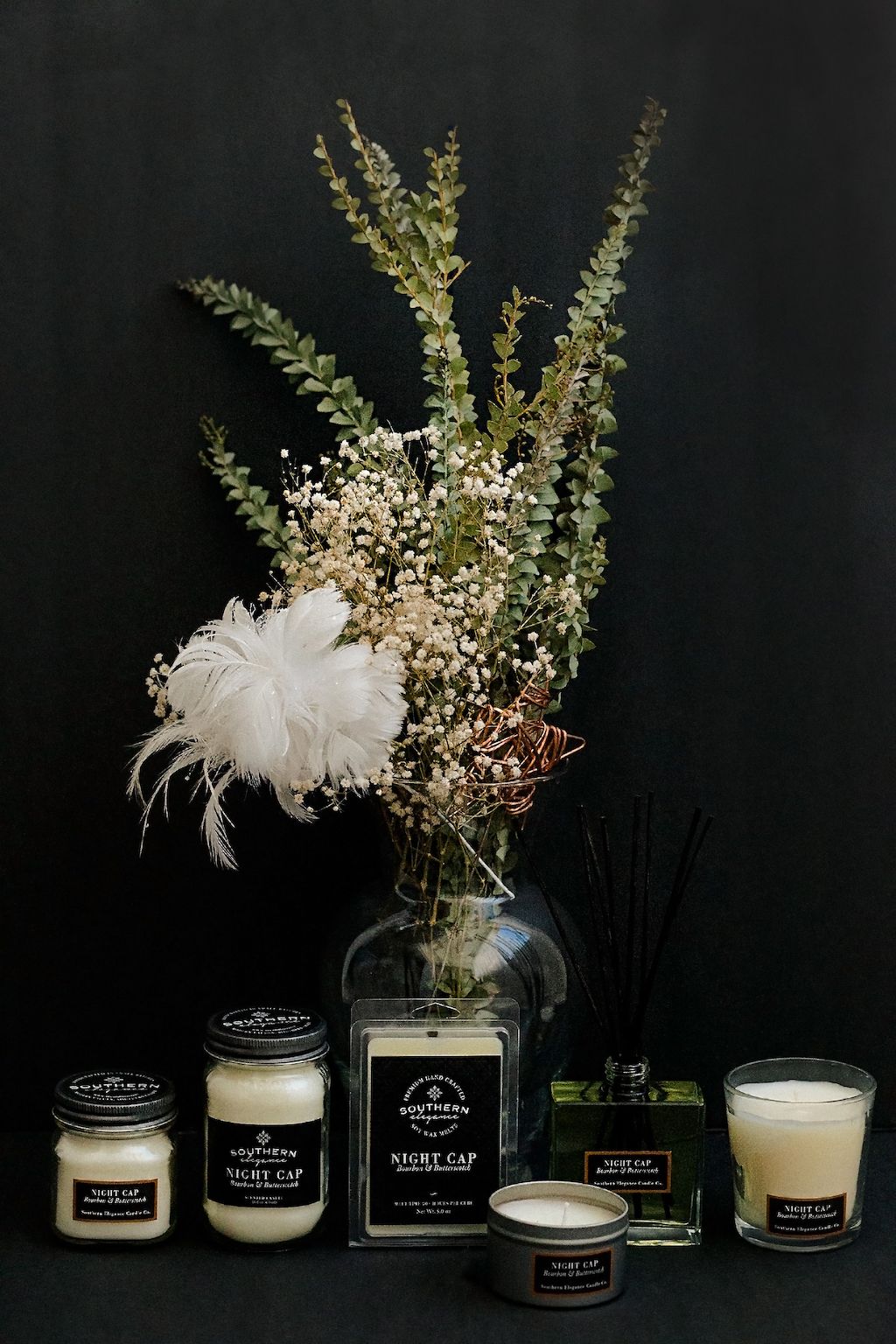 Bundle of soy wax candles with a vase of greenery and white feathers on a black background