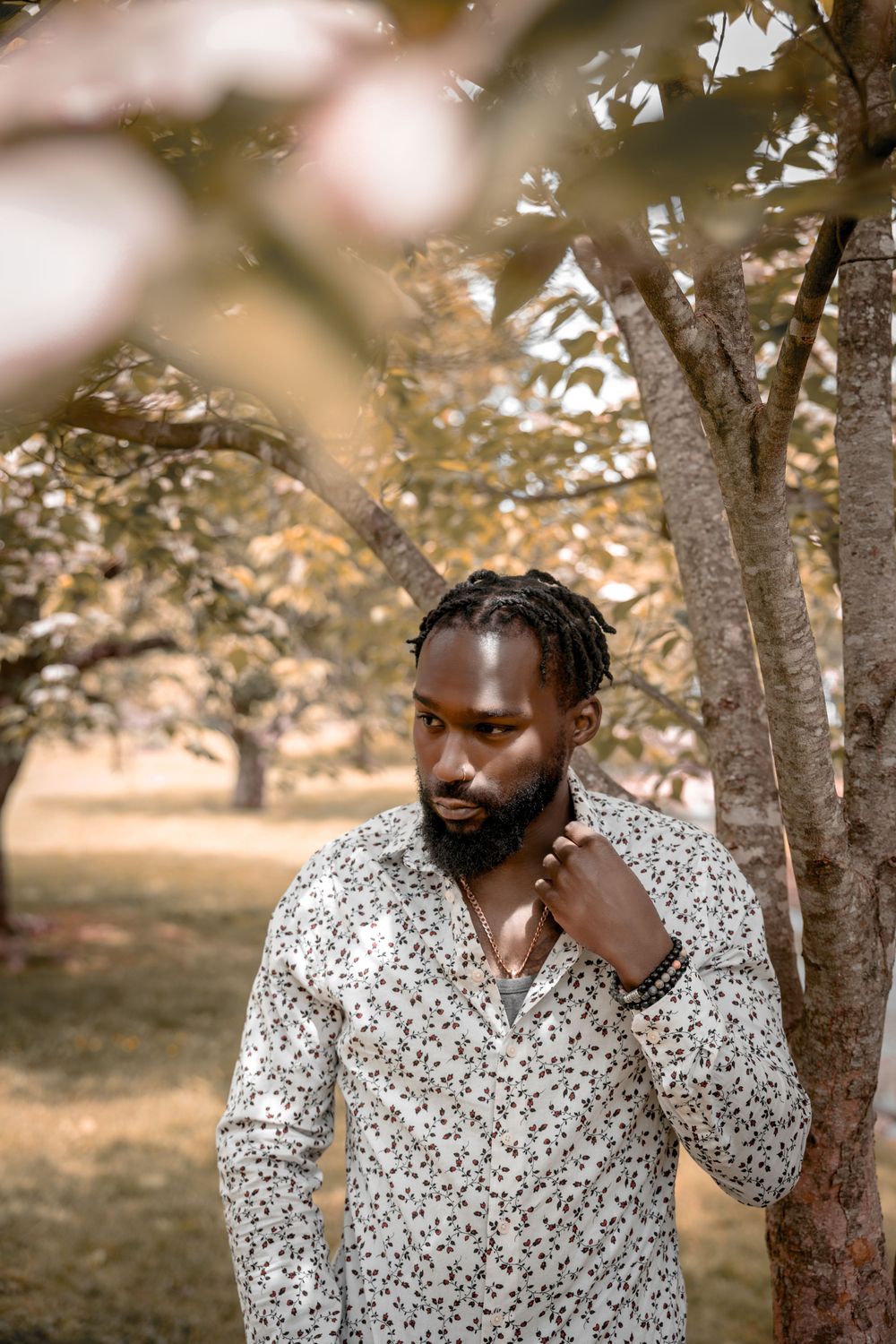 Male model wearing a floral print shirt poses thoughtfully in an outdoor setting with autumn trees in the background.