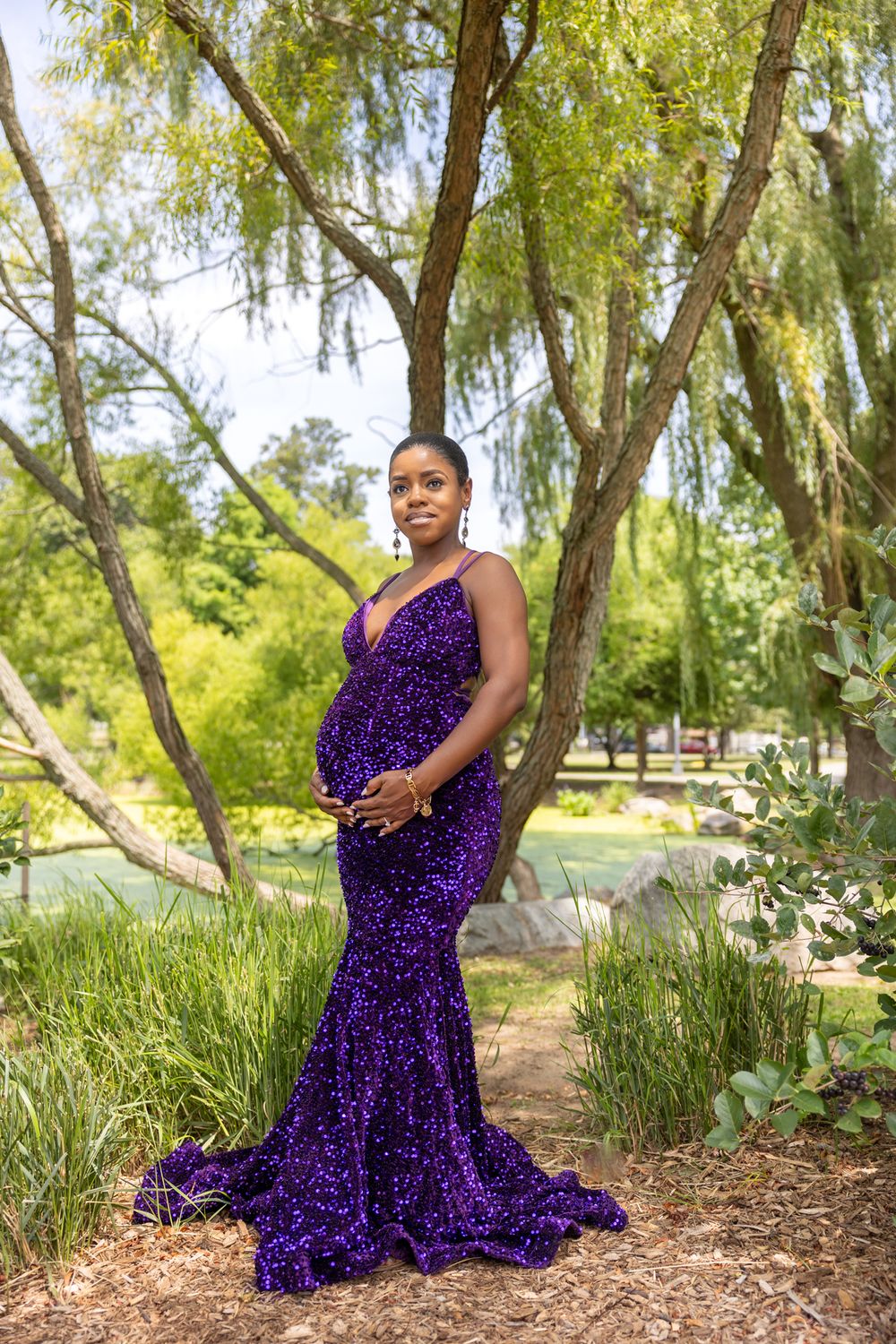 Elegant maternity photo featuring a sparkly purple mermaid gown in a lush garden setting with willow trees.