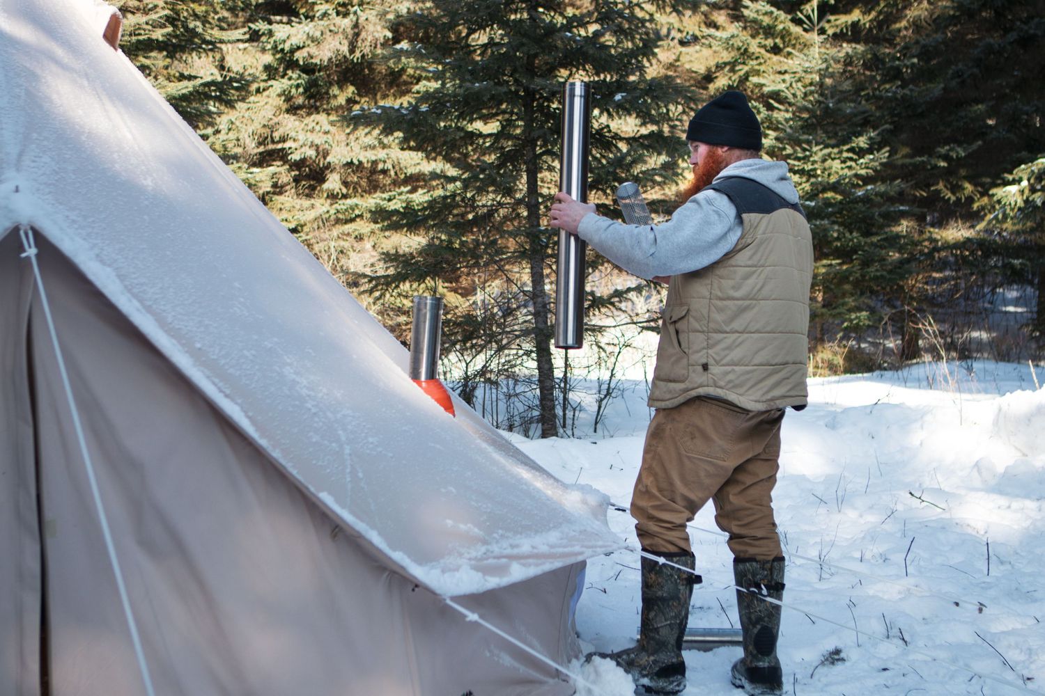 Person installing stove pipe through tent wall in snowy winter setting.
