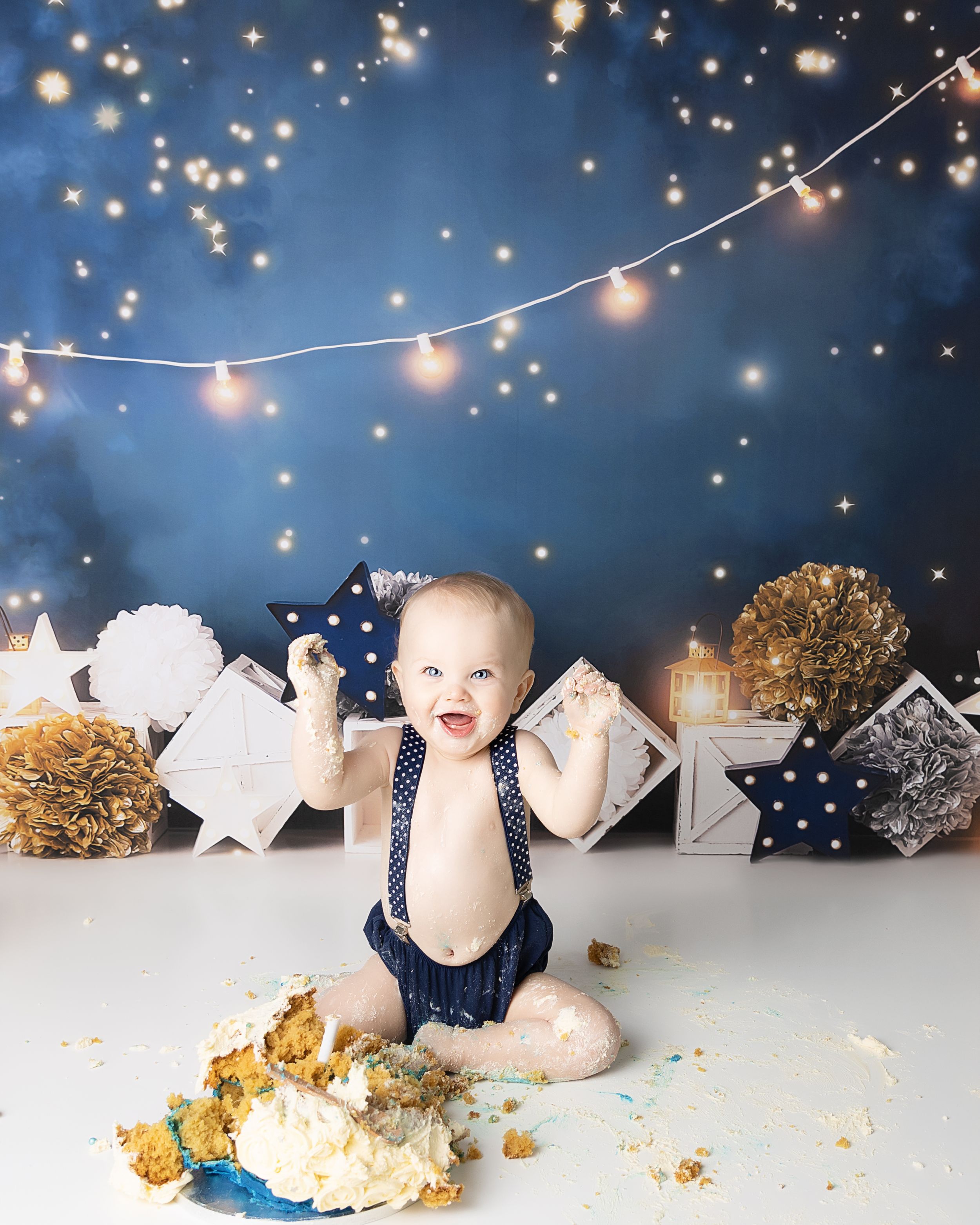 A baby in navy overalls sits on a white backdrop with sparkly string lights and decorative elements during a cake smash photo shoot.
