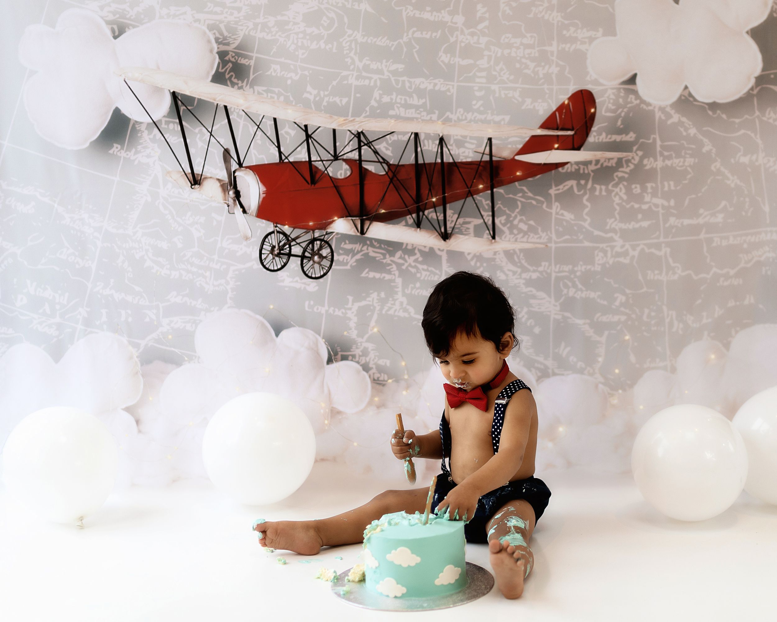 A red vintage airplane decoration hangs above a child having a birthday celebration with cake and white balloon clouds.
