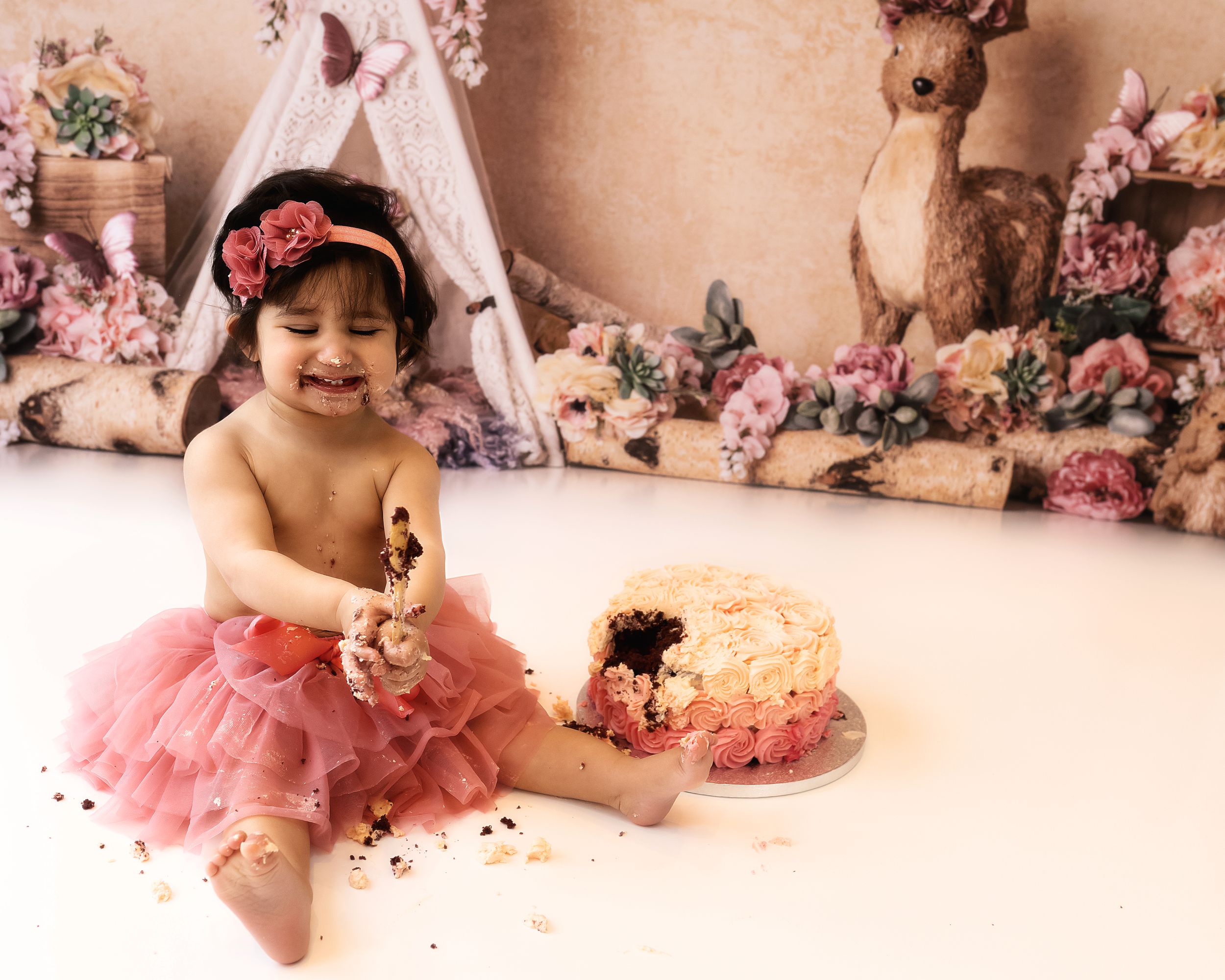 A baby wearing a pink tutu dress sits next to a decorated cake in a floral themed room with a teepee and plush deer decor.