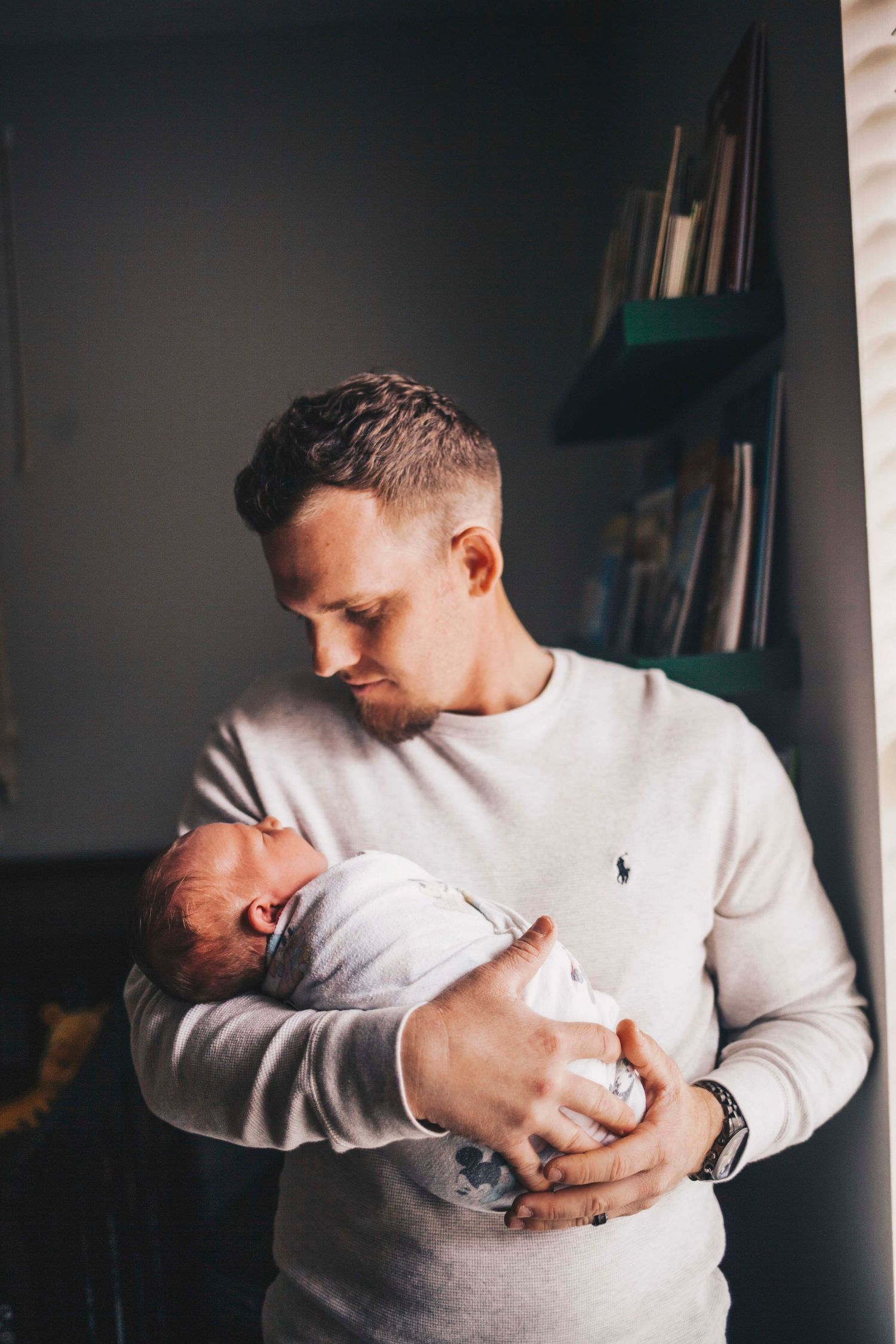 New father in white sweater tenderly cradling sleeping newborn baby near window indoors.
