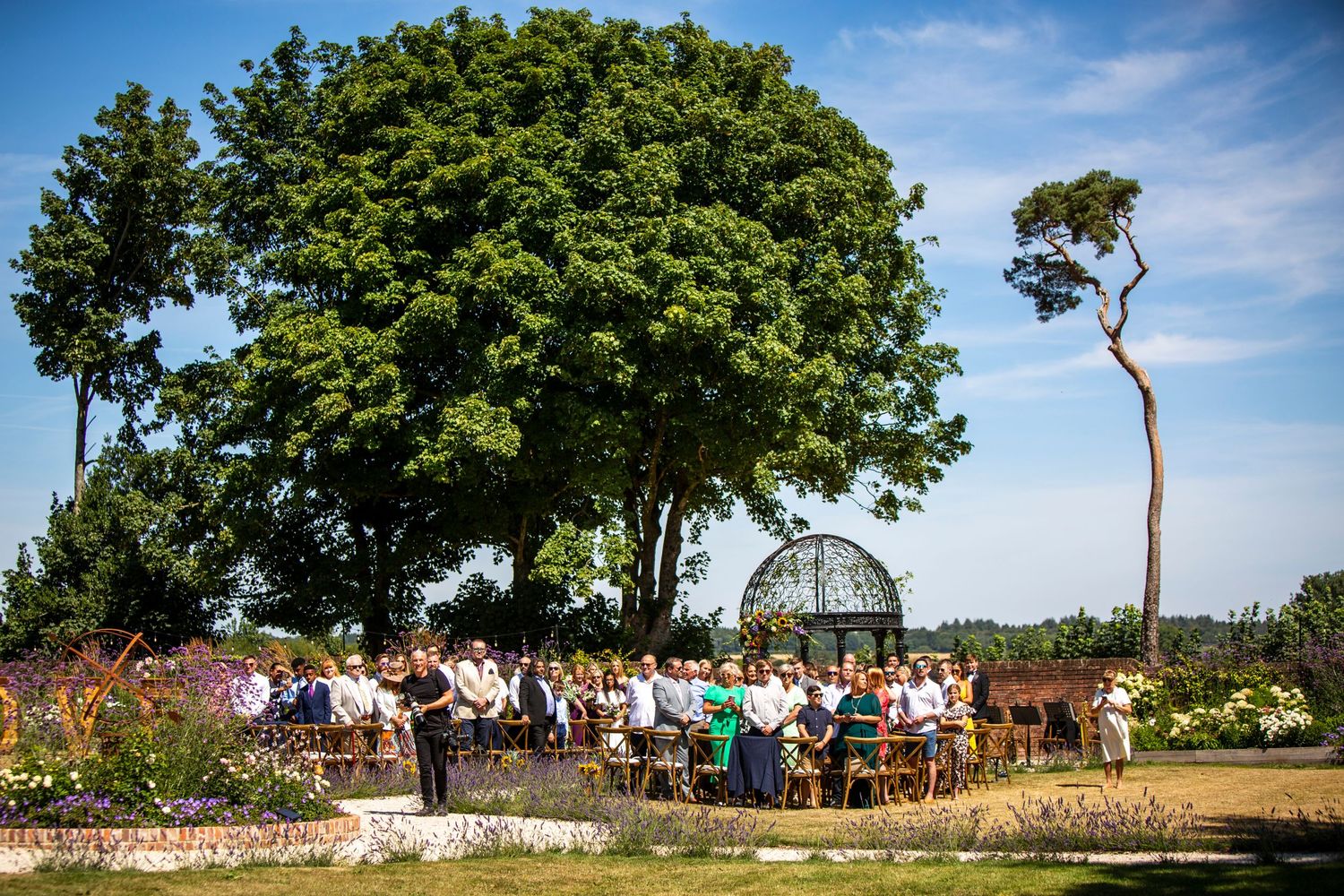 Outdoor wedding ceremony setup with rows of chairs under trees and blue sky on a sunny day.