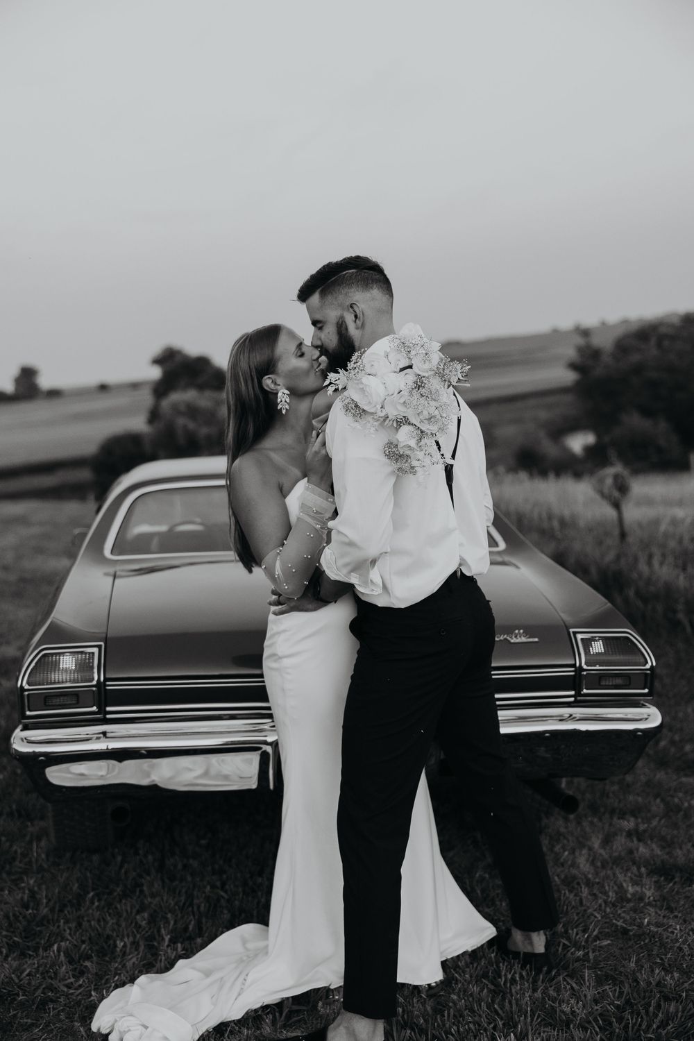 A romantic couple embraces by a vintage car in a rustic field during sunset.