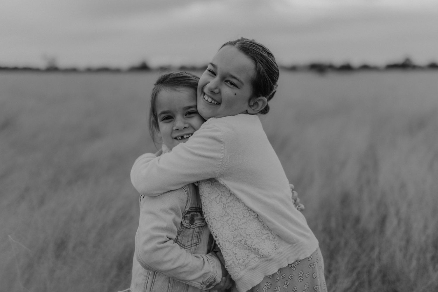 Two children embrace in a black and white portrait in a grassy field.