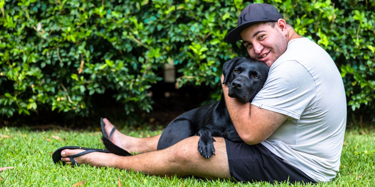 This photo has a 20 year old male sitting on the grass outdoors cuddling his black Labrador, guide dogJonah.
