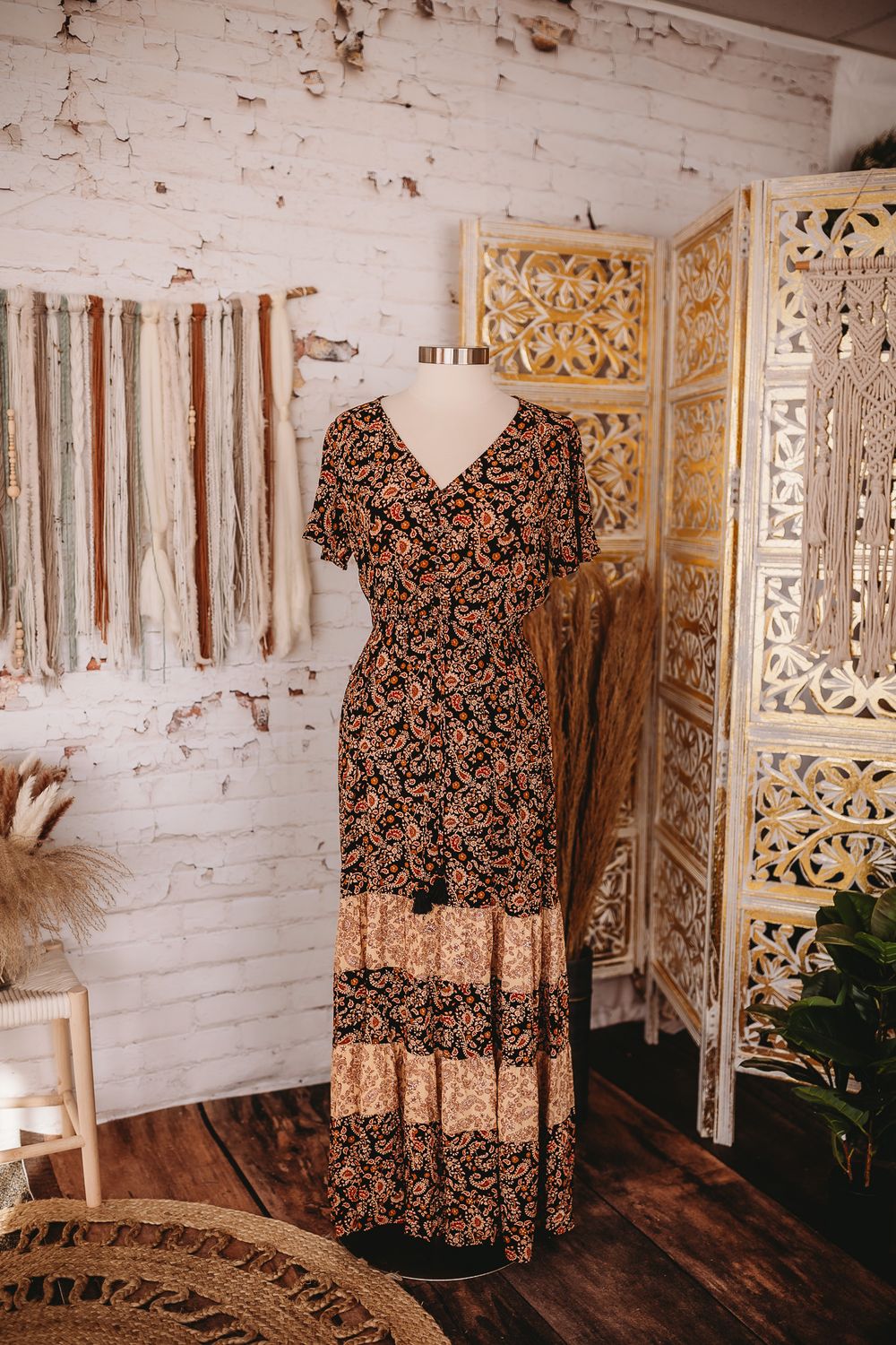 Black floral print tiered maxi dress on mannequin in bohemian studio with white brick wall decor.