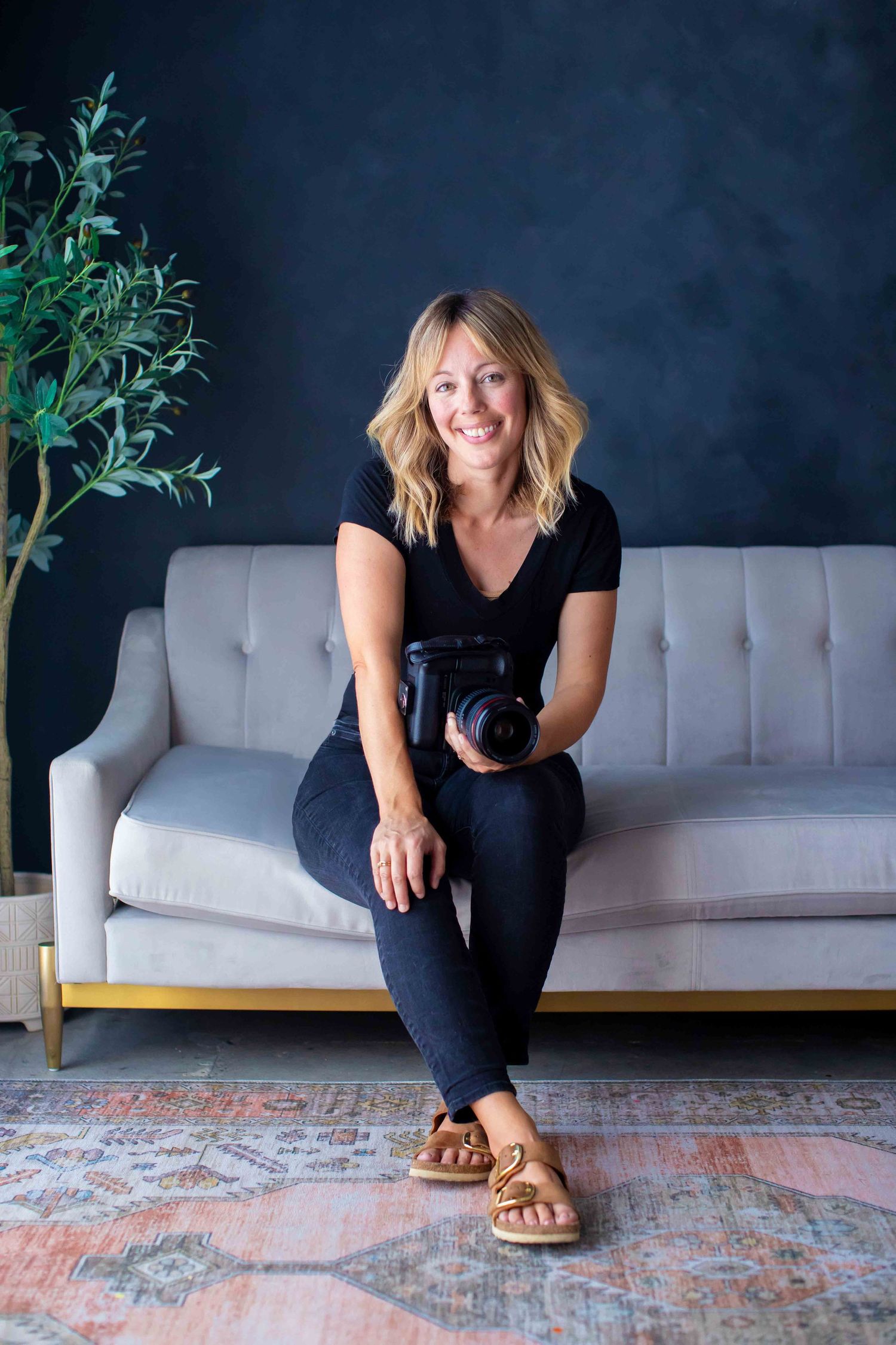 Woman in black outfit sits casually on carpet near gray couch and greenery.