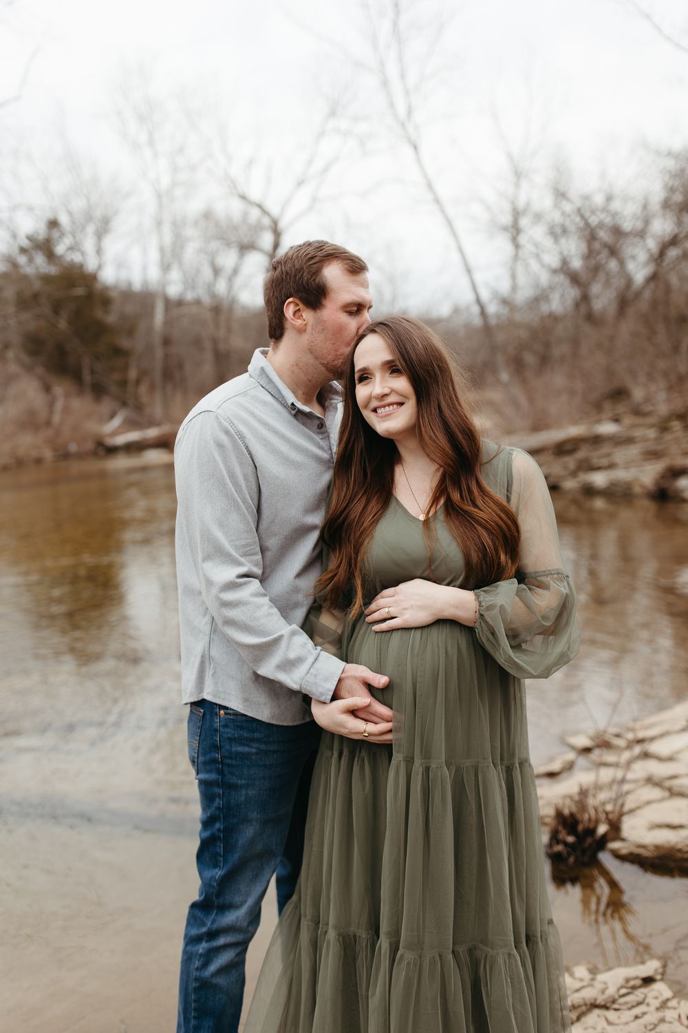Couple sharing tender moment by riverside during maternity photoshoot.