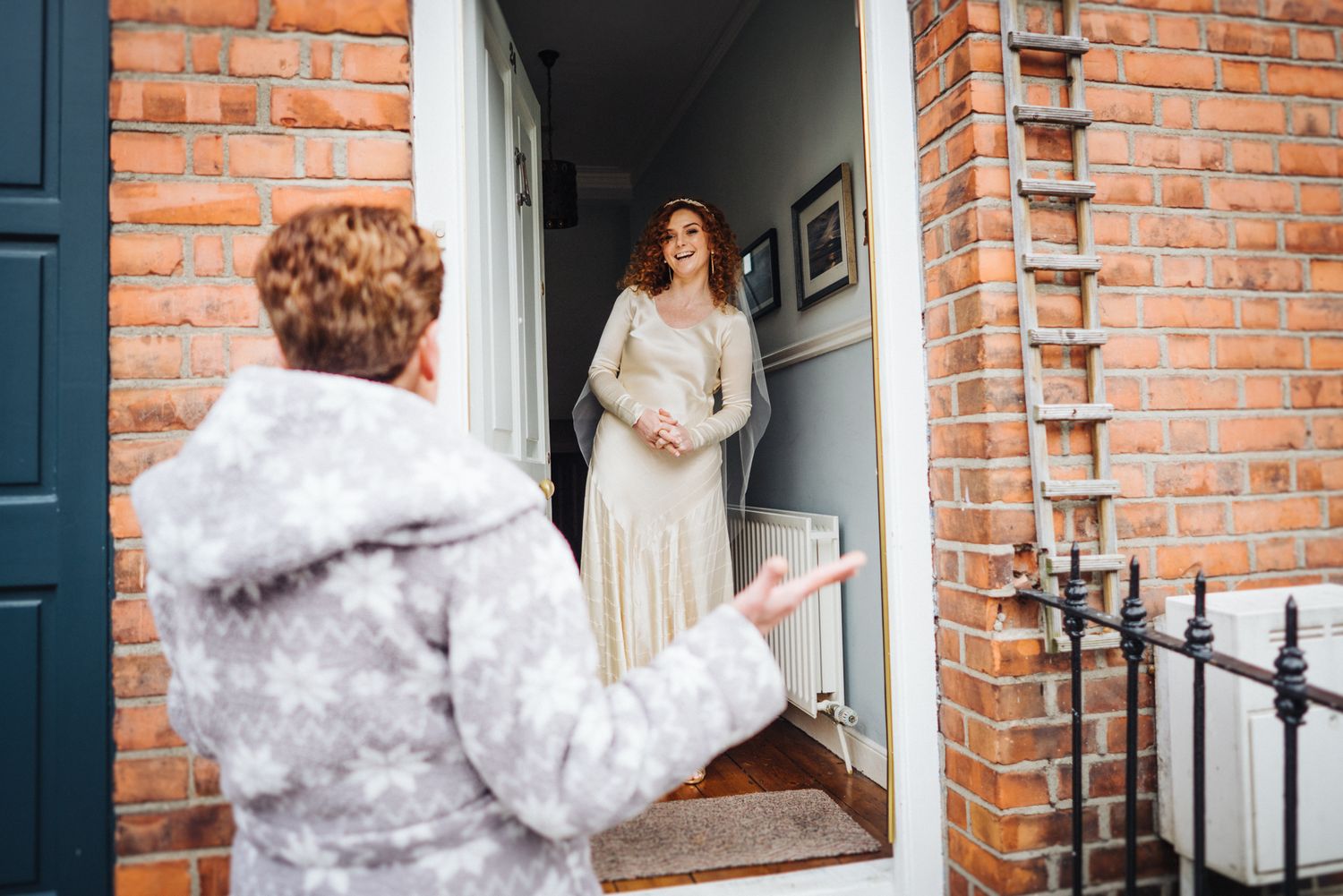 Winter wedding scene with person in coat greeting someone in elegant dress at brick doorway.