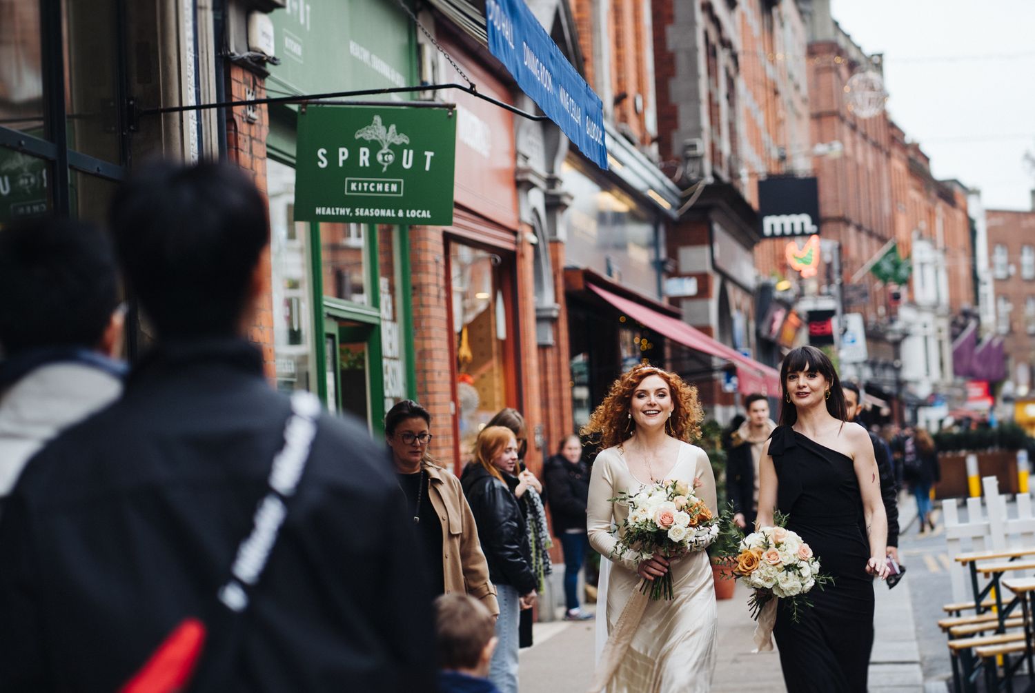 A wedding party walks down a busy urban street past shops and storefronts.