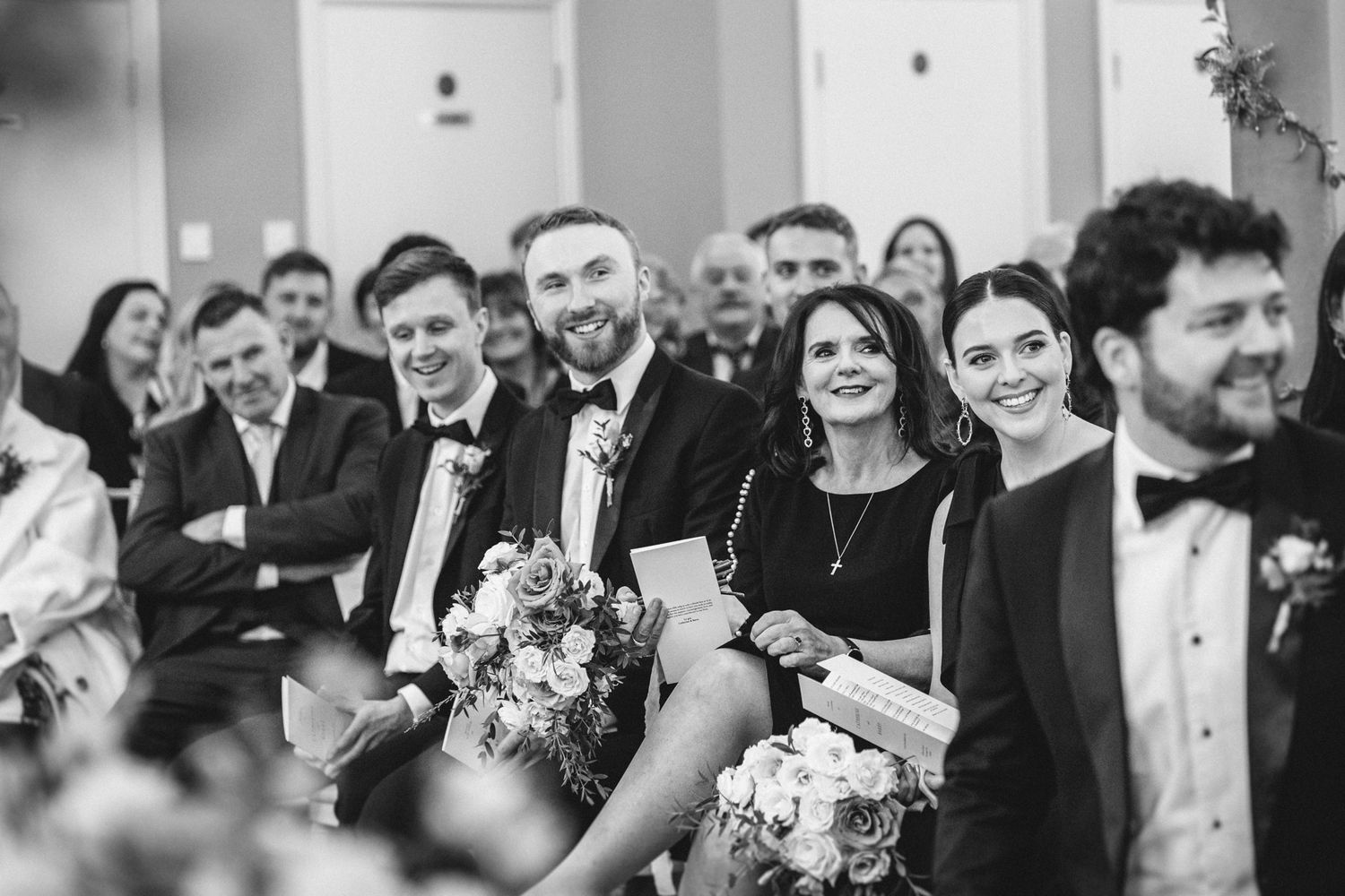 Wedding guests smile and react during an indoor ceremony captured in black and white.