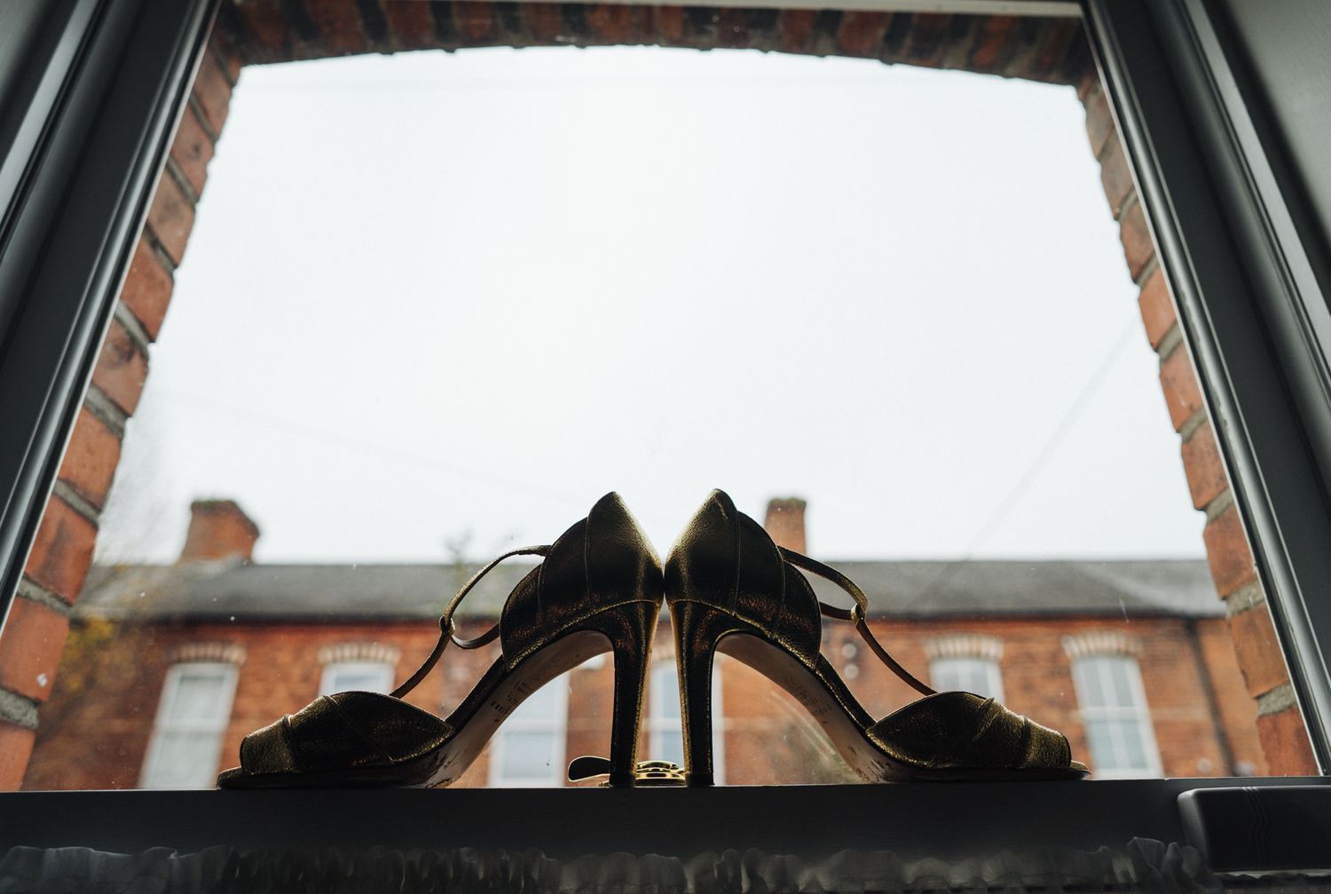 Pair of elegant black high heels silhouetted against a window with brick building in background.