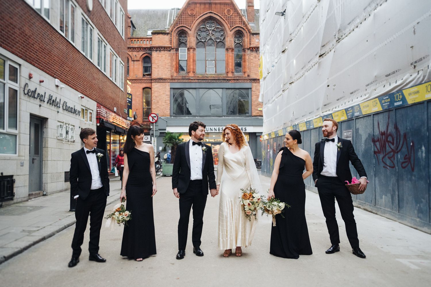 Wedding party poses together on urban street with historic red brick building.
