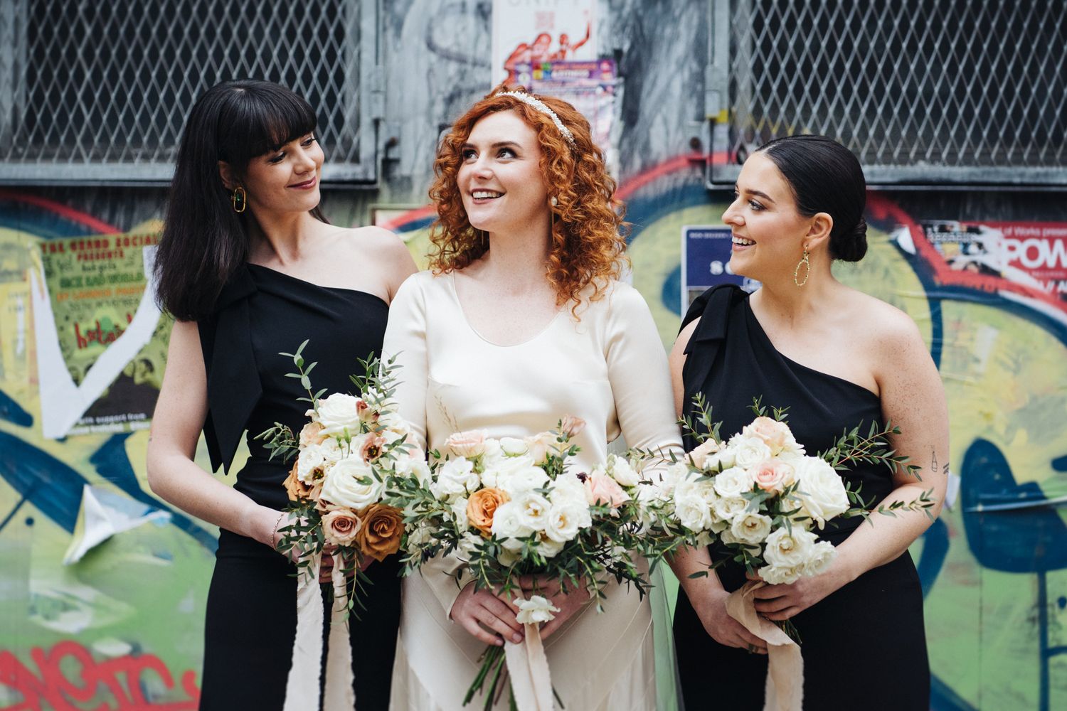 Three attendants in black dresses hold floral bouquets against graffiti wall.