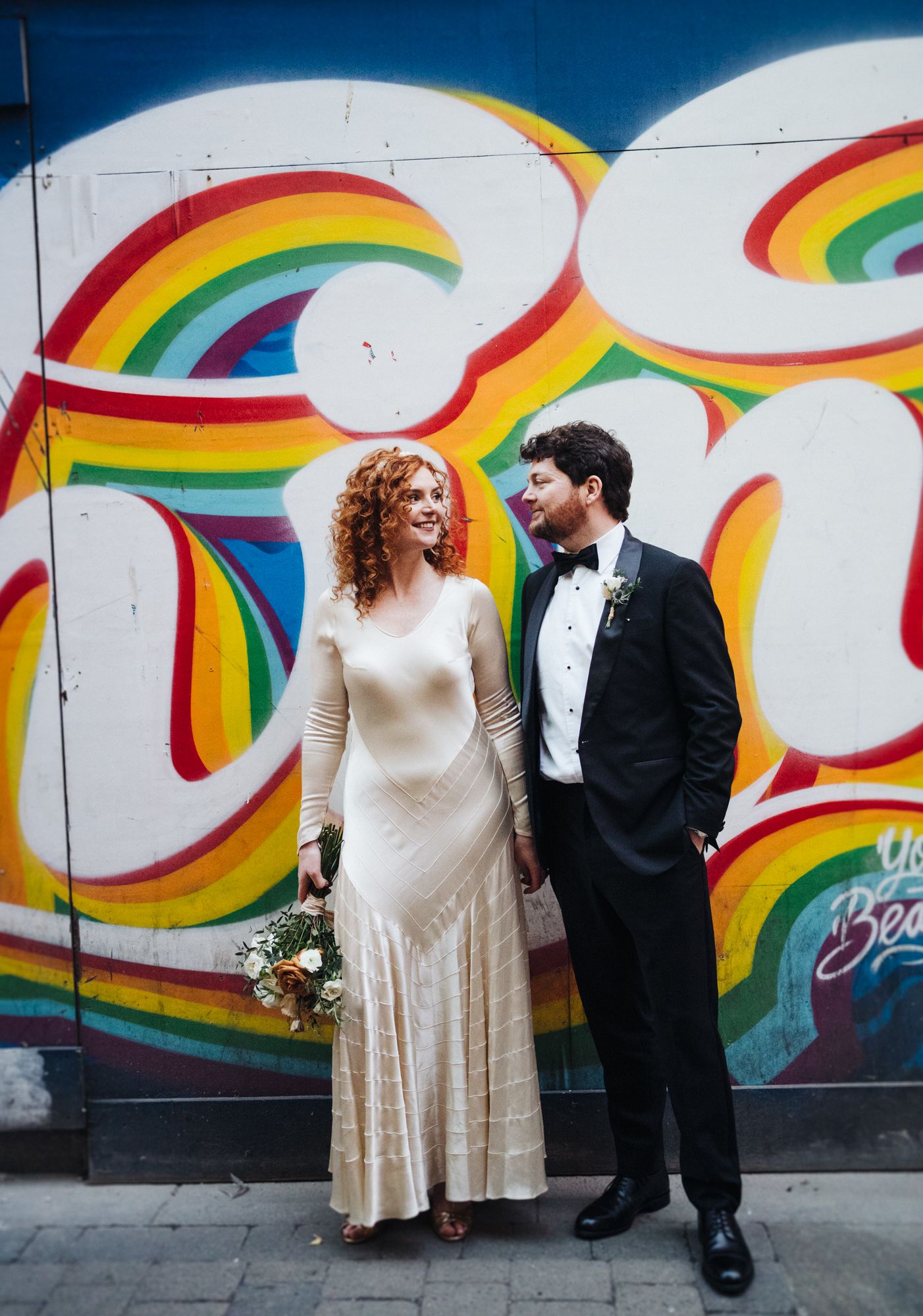 Elegant couple stands before colorful rainbow mural in wedding attire.