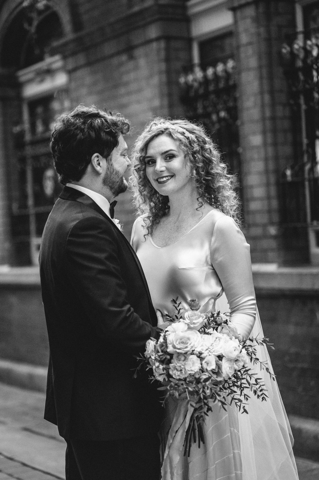 A black and white wedding portrait of a couple sharing a romantic moment against an elegant building facade.
