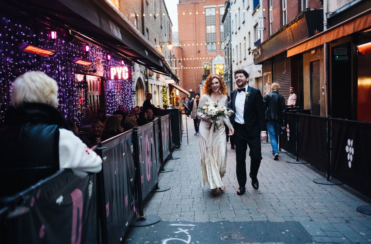 A couple walking down a city alley illuminated by decorative string lights and neon signs at dusk.