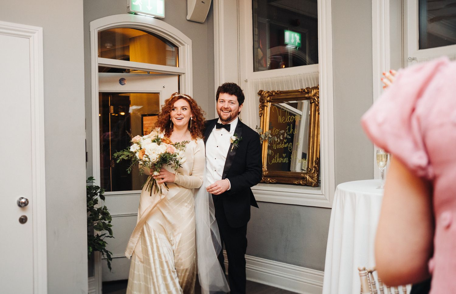 A couple stands elegantly in a bright interior doorway with ornate decor.