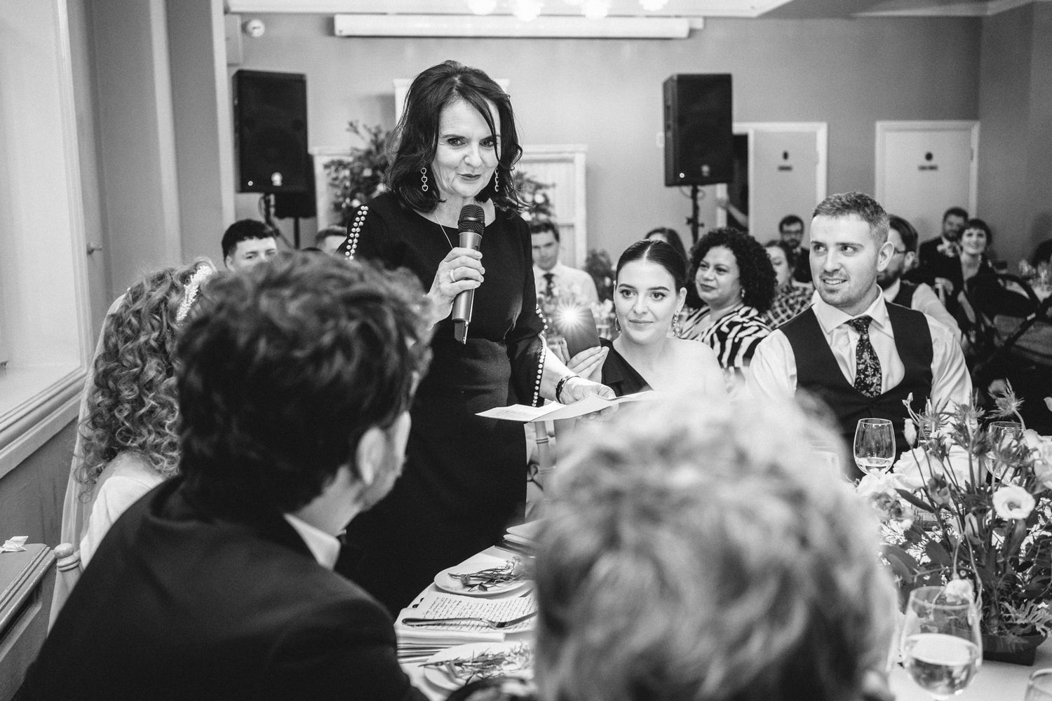 A candid moment at an elegant dinner reception as guests gather around decorated tables while someone makes a speech.