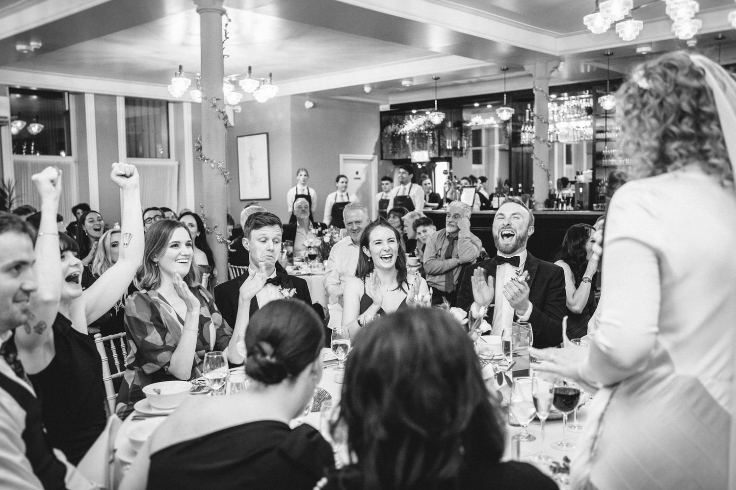 Wedding reception guests gathered in a well-lit dining room react enthusiastically to a speech.