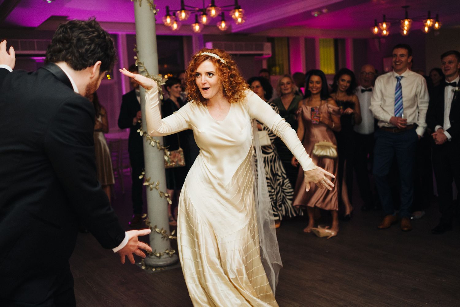 Bride in white dress dancing energetically at wedding reception with purple lighting.