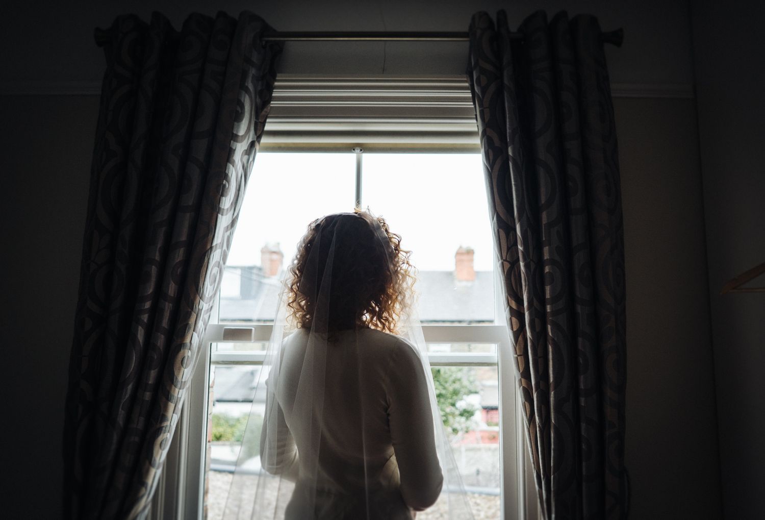 Silhouette of person in white dress looking out window with dark curtains.